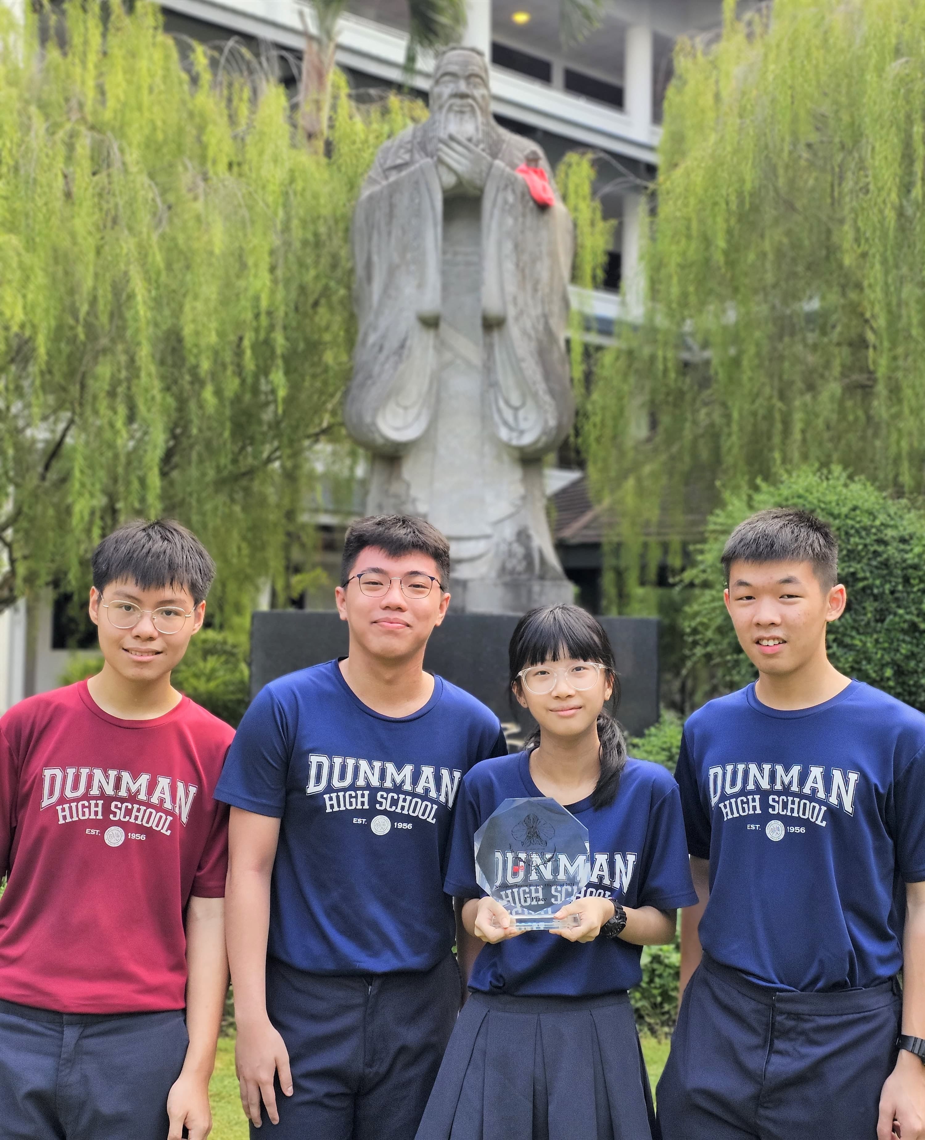 Four Dunman High School students pose before a large stone statue, one girl holding a crystal trophy.