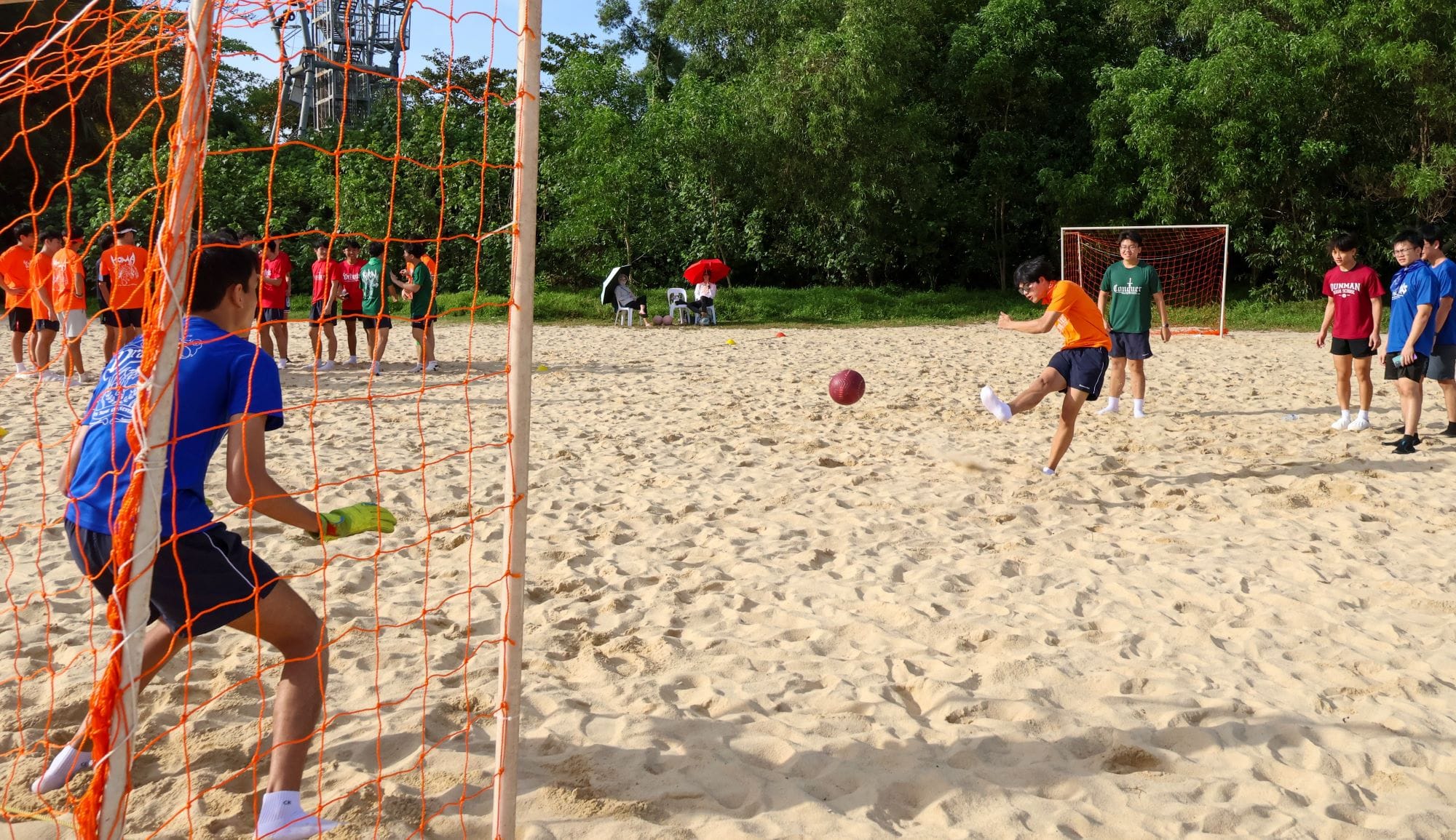 Young people playing beach soccer on sand, with a player kicking toward an orange net goal.