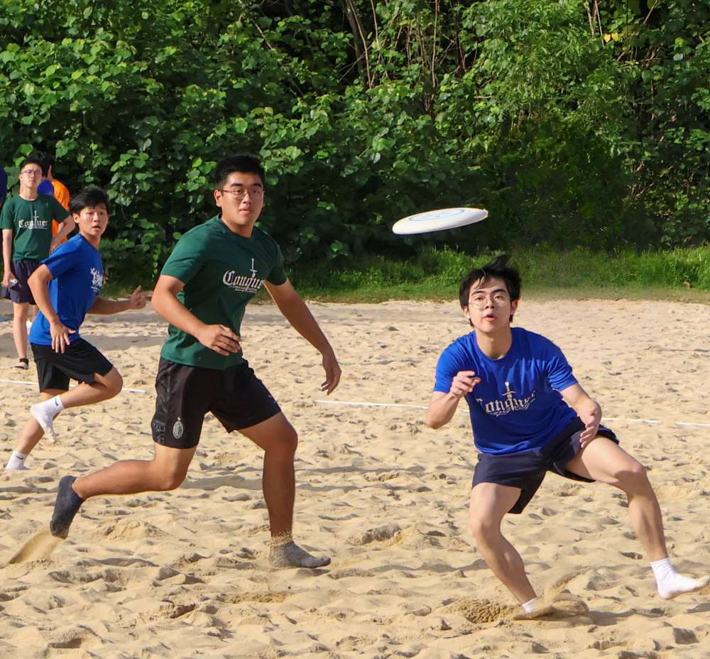 Several young people play frisbee on a sandy beach court, with a white disc flying through the air.
