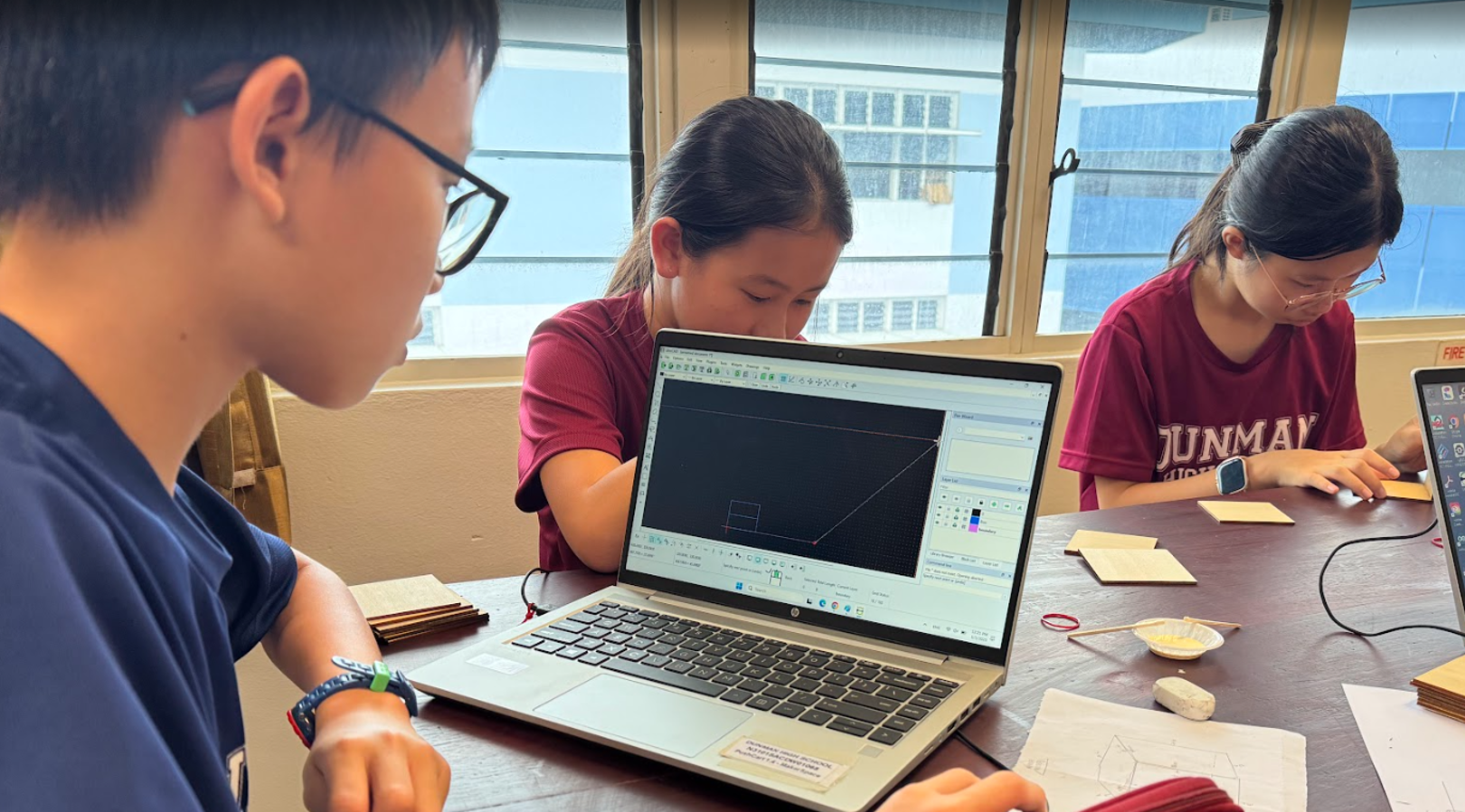 Three students work at a table, with two looking at a laptop running CAD design software.
