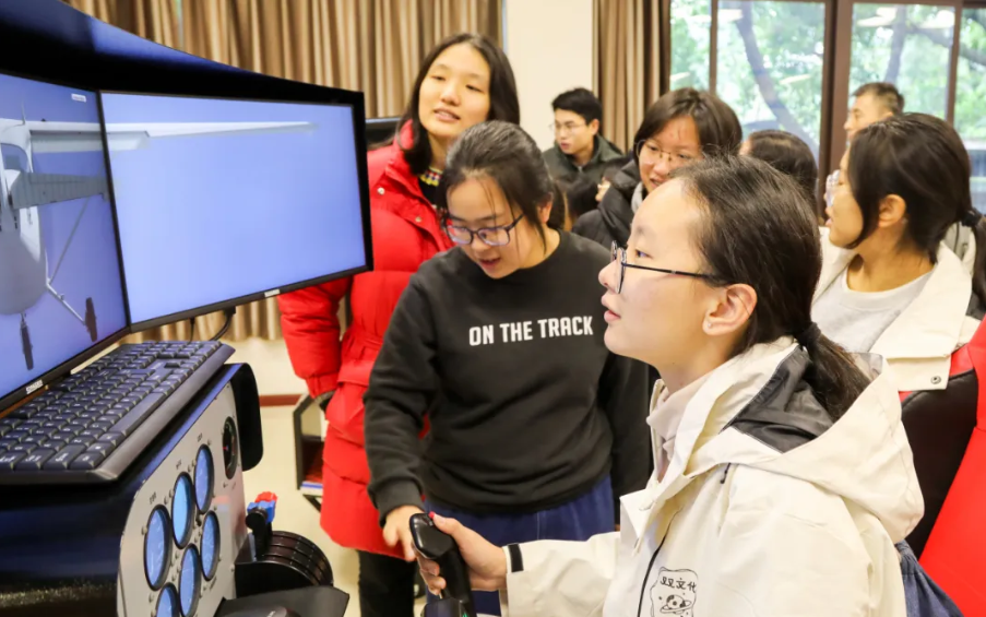 A young woman uses a flight simulator joystick while a group of students watches the airplane displayed on a curved monitor.