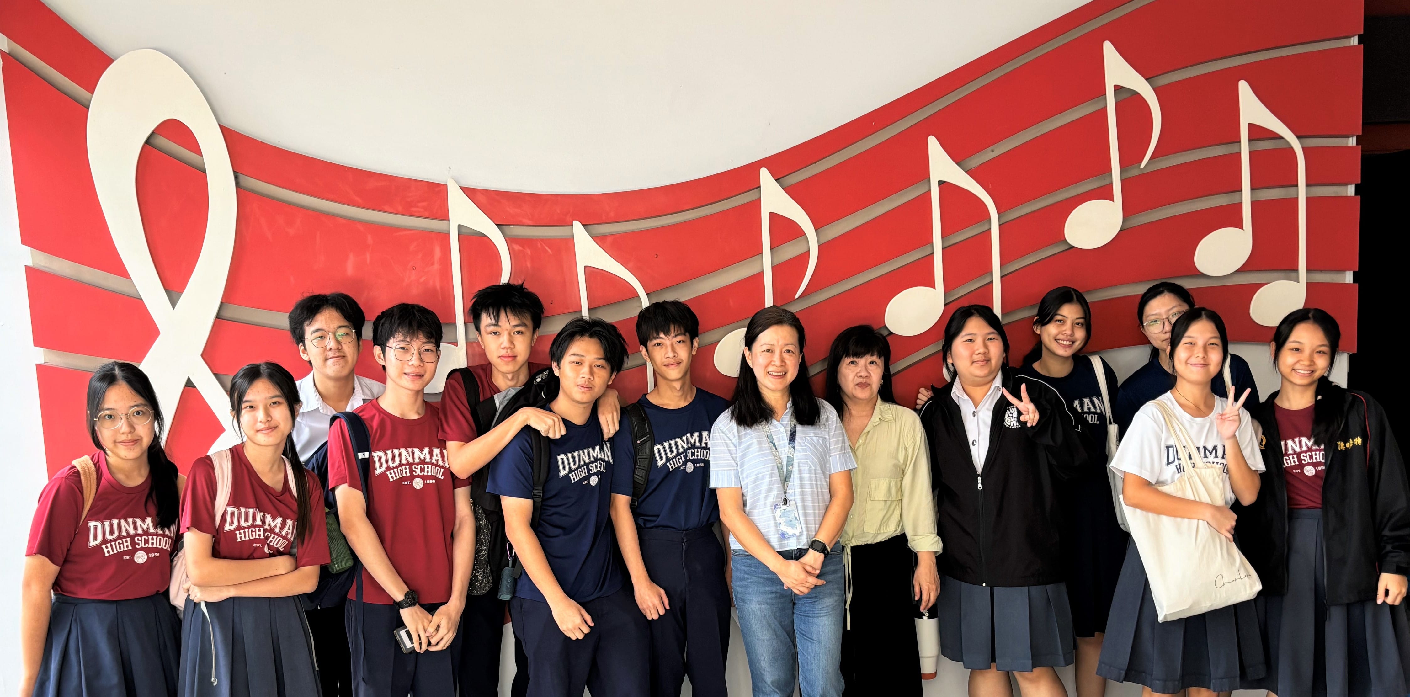 Group of Dunman High School students and adults posing in front of a red wall with large white musical notes.