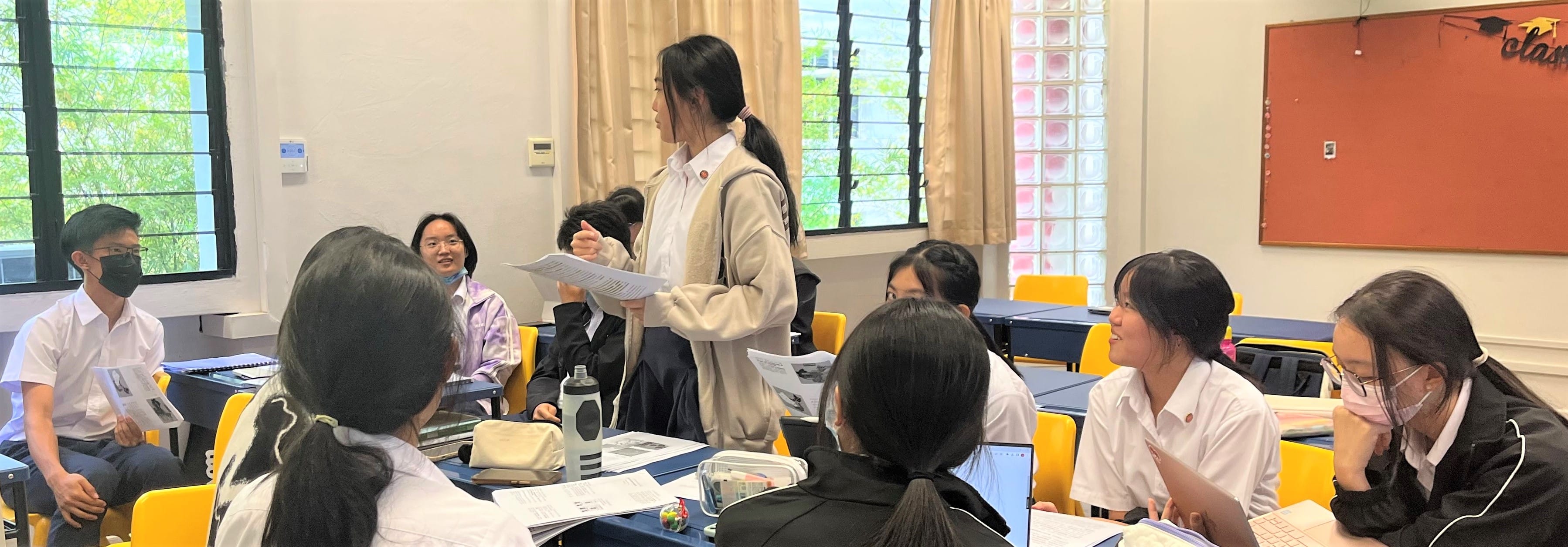 A student stands holding papers while classmates sit at desks with books in a bright classroom.