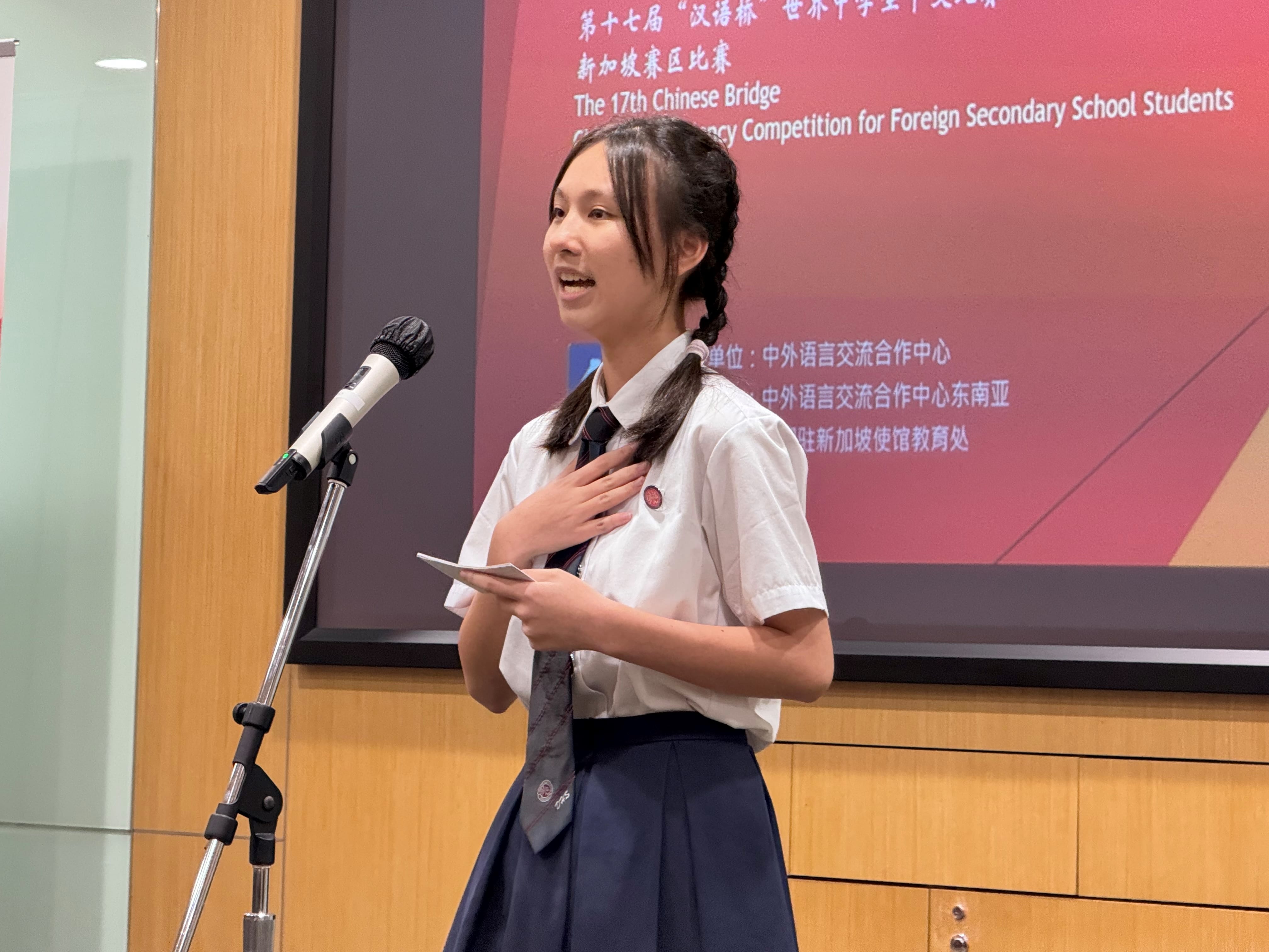 Student in school uniform speaks at a microphone, holding note cards, with a competition banner behind her.