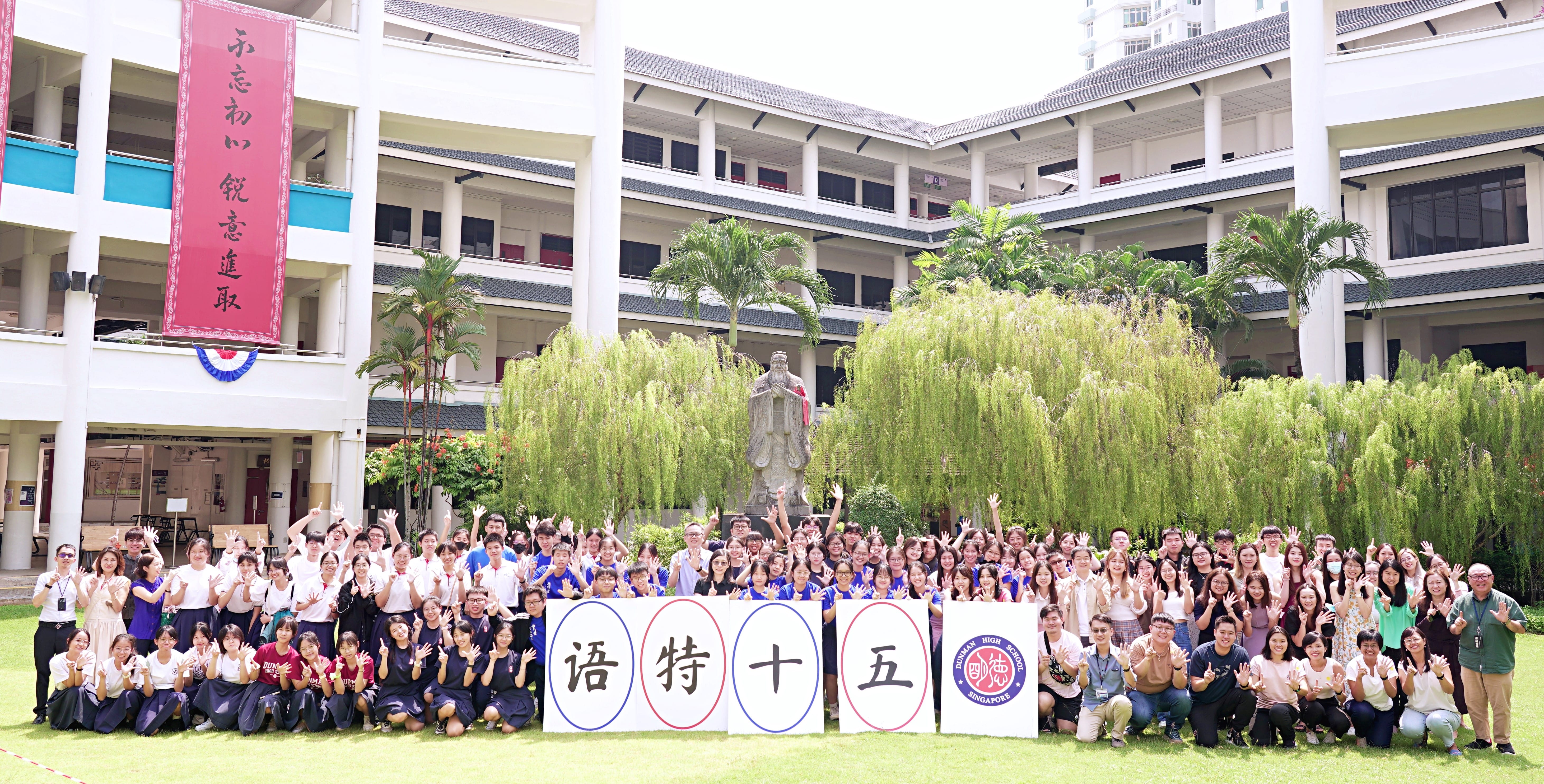 Large group posing in a school courtyard, holding Chinese character boards; statue and trees behind.