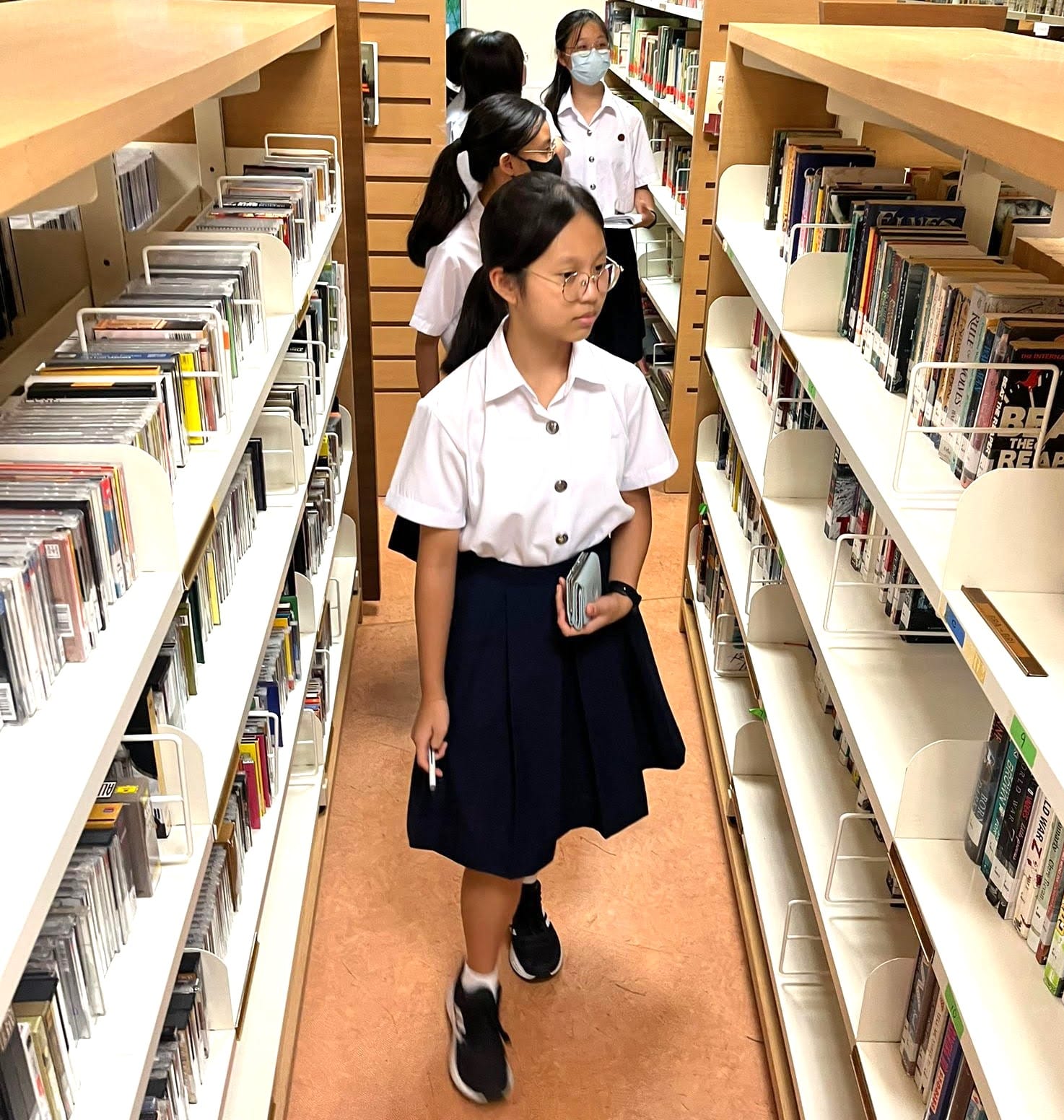 Several school girls in white shirts and navy skirts walk through a library aisle lined with bookshelves.