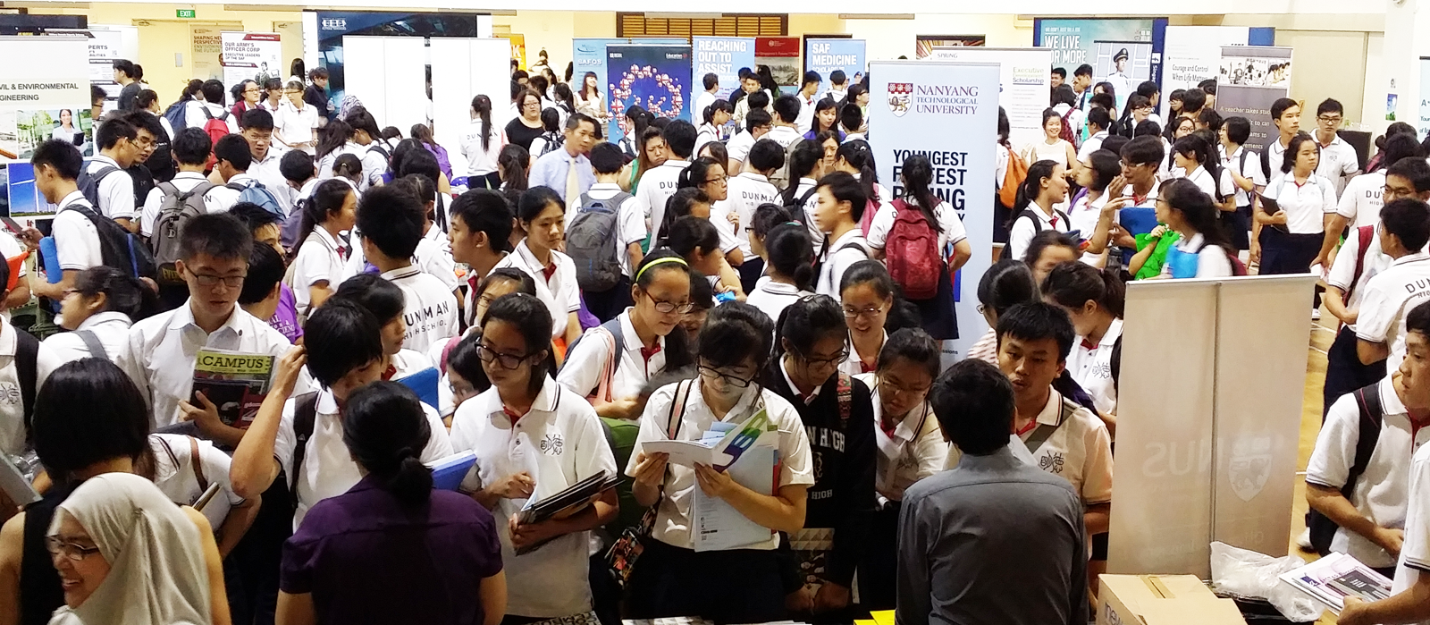 Students in white uniforms browse brochures at a crowded education fair with Nanyang Technological University banners visible.