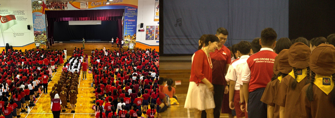 Students in red uniforms fill a school hall facing a stage, while adults speak with uniformed students nearby.