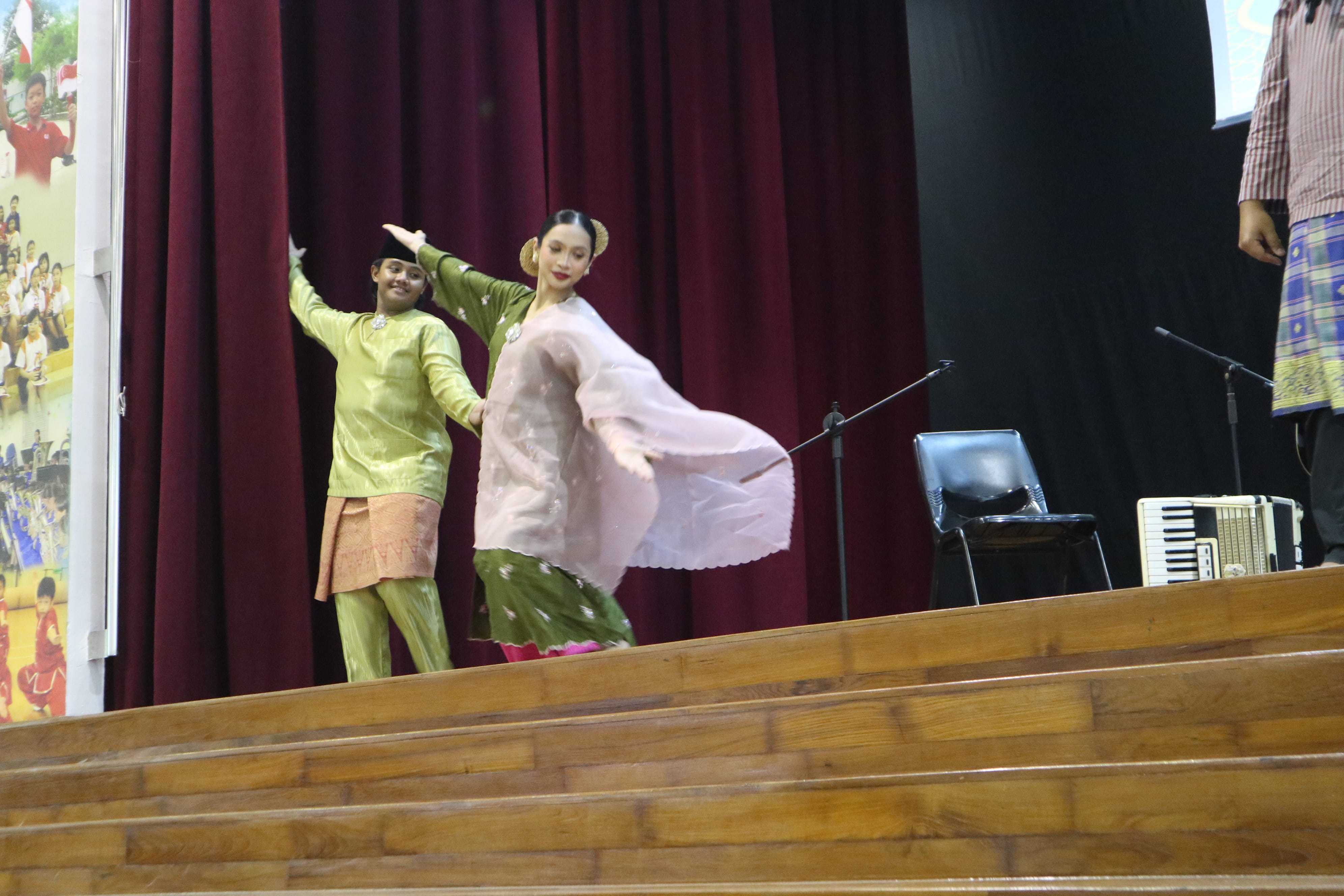 Two dancers in traditional dress pose on stage before maroon curtains; microphones, chair, and accordion nearby.