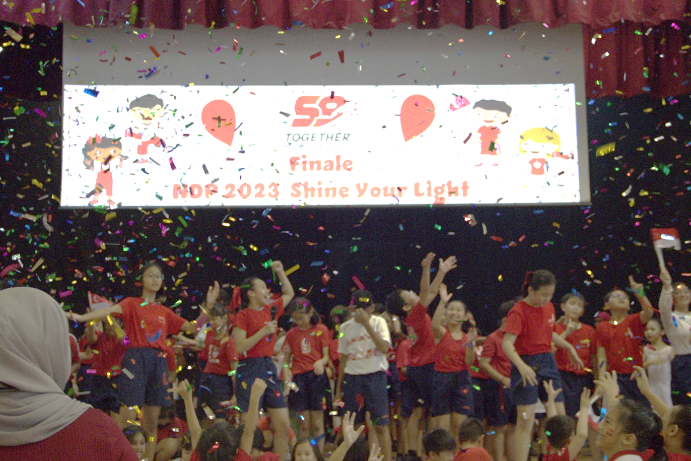 Children in red shirts celebrate on stage with colorful confetti falling, and a screen reading "Finale NDP 2023 Shine Your Light."