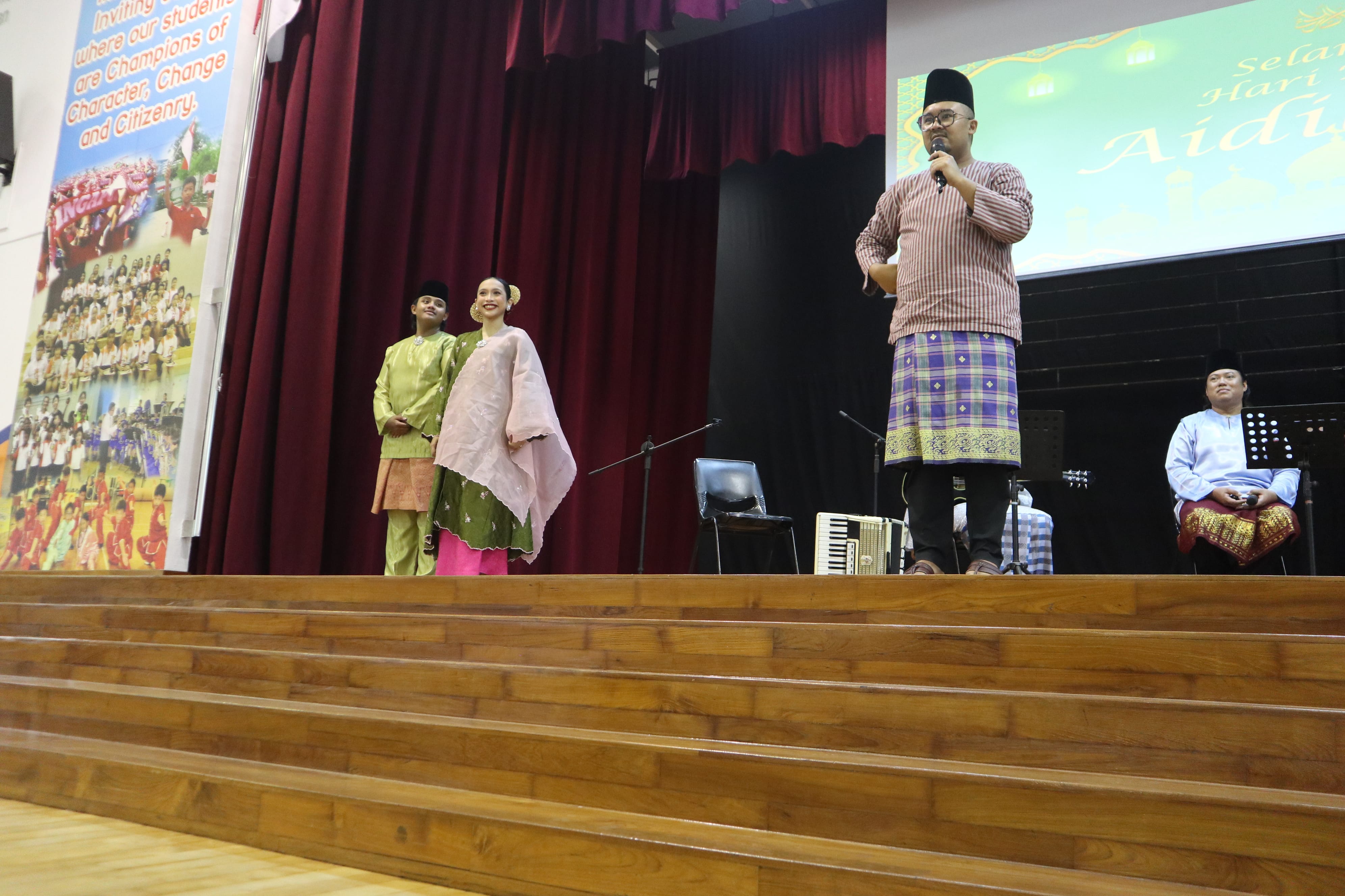 Man in traditional attire speaks into microphone on stage; two in matching outfits pose near red curtains.