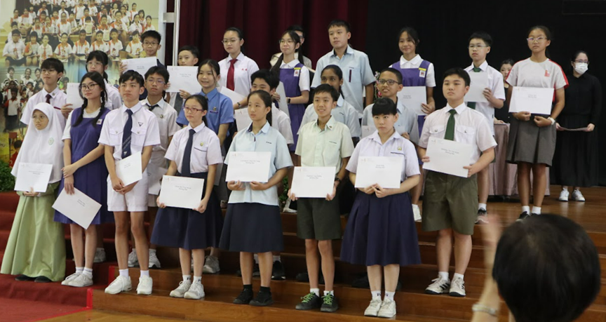Students in school uniforms stand on a stage holding certificates during an award ceremony.