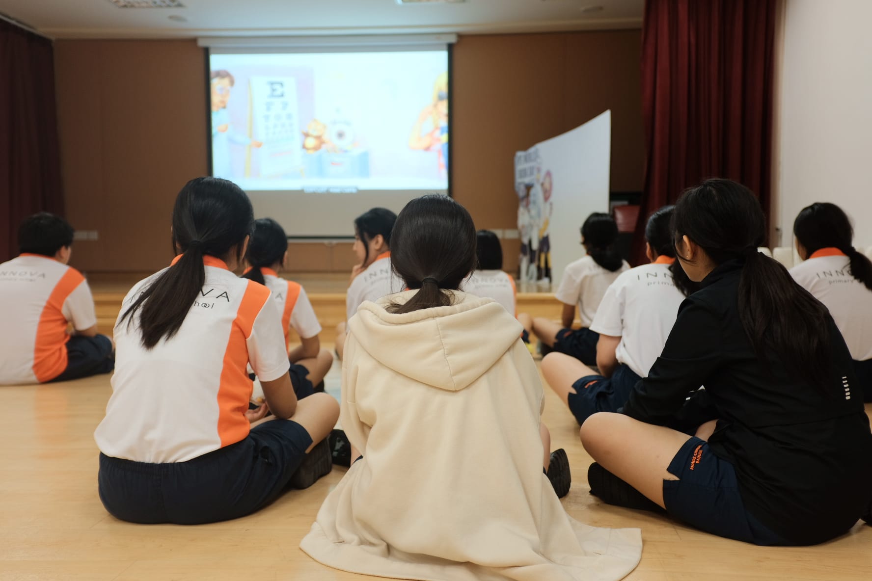 Students in school uniforms sit on a wooden floor watching a projected animated presentation on a screen.