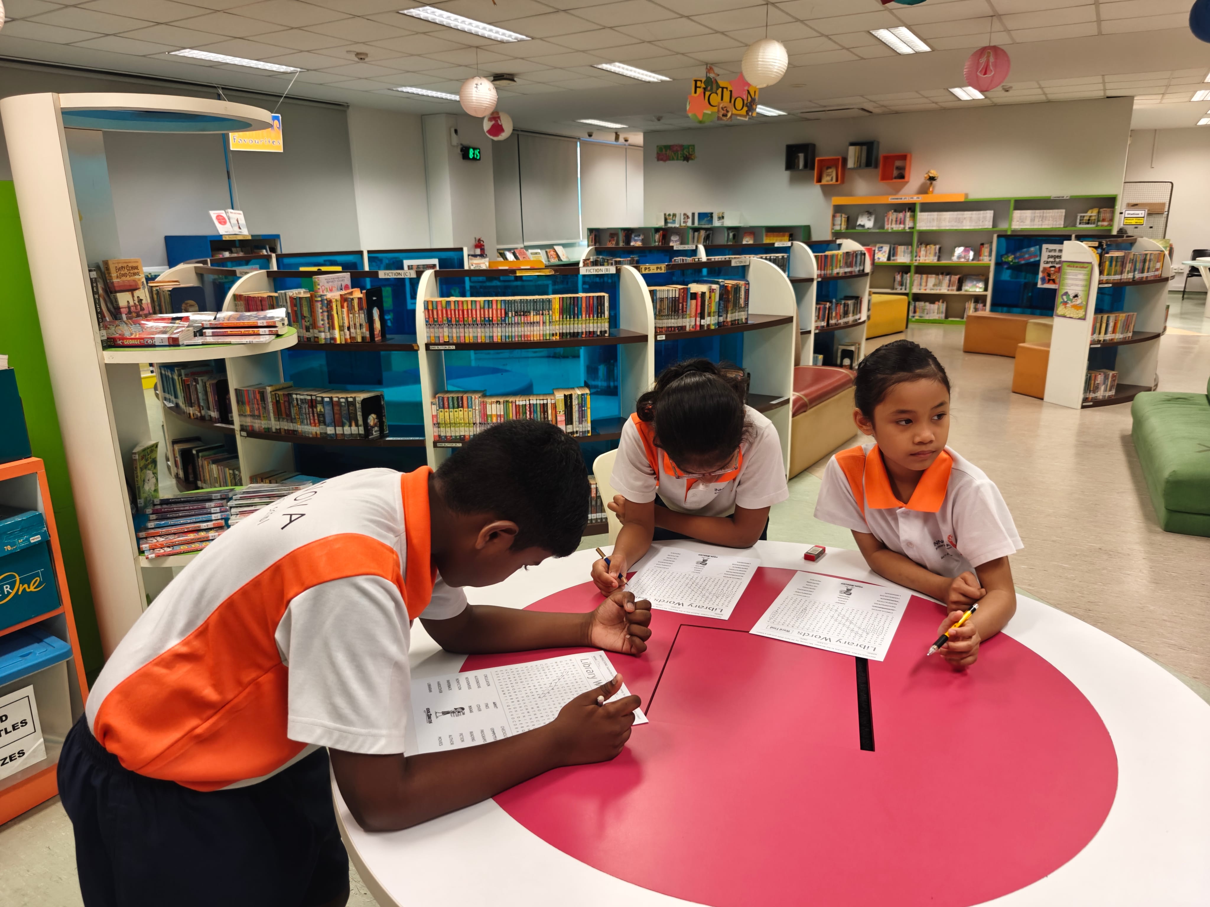 Three children in orange and white uniforms write on worksheets at a round pink table in a school library.