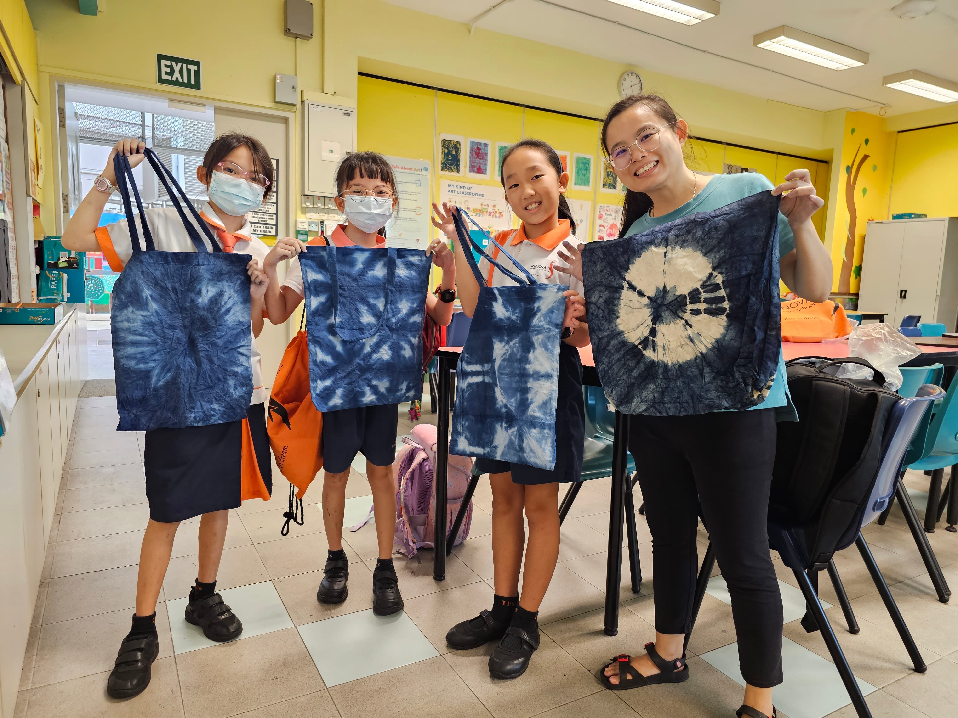 Three children and an adult smile while holding up blue and white tie-dyed tote bags in a school art classroom.