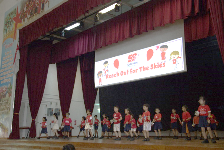 Young children in red and white outfits perform on a stage with a banner reading "Reach Out for The Skies."