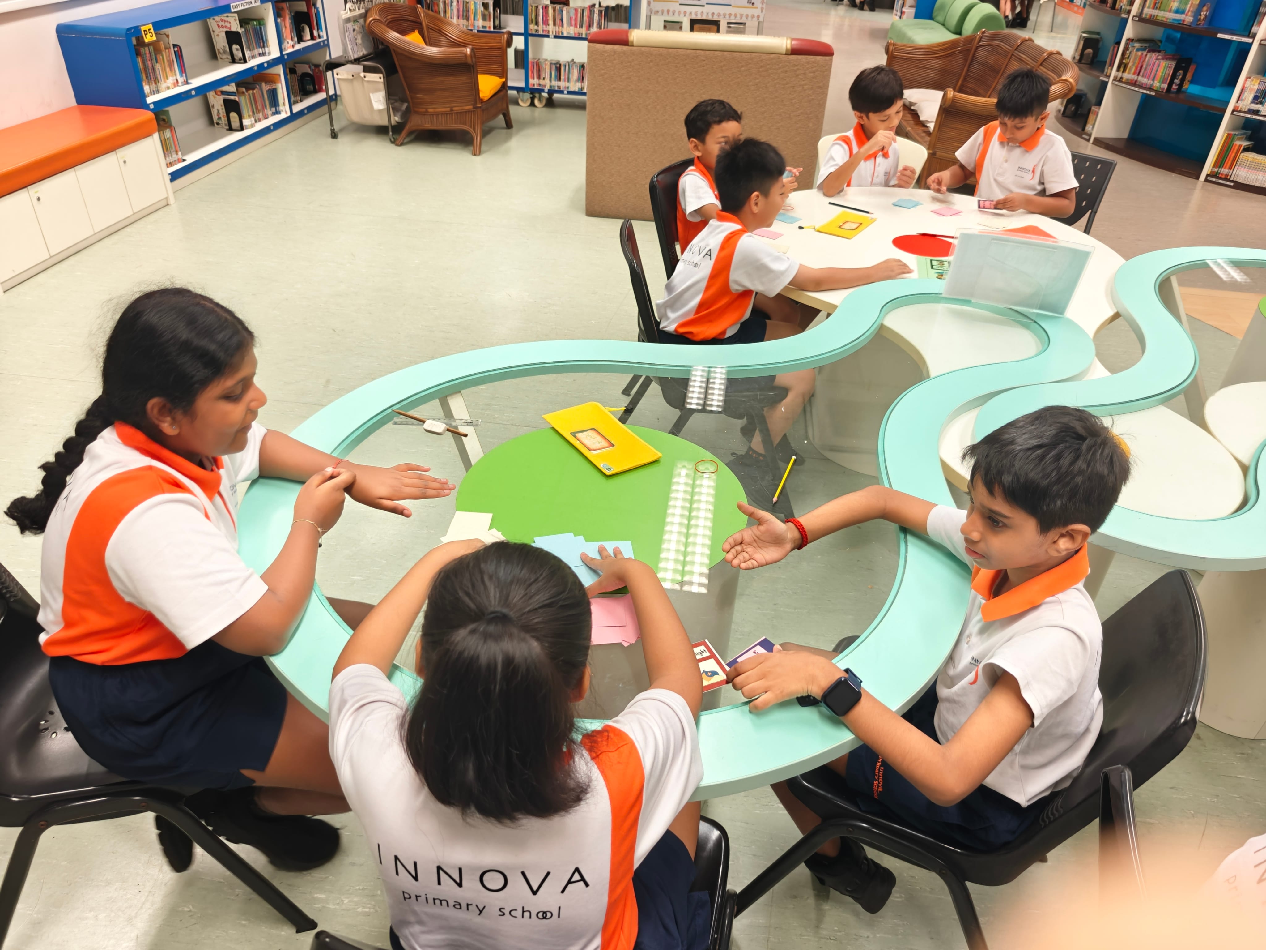 Children in Innova Primary School uniforms sit around round tables in a library, working on group activities.