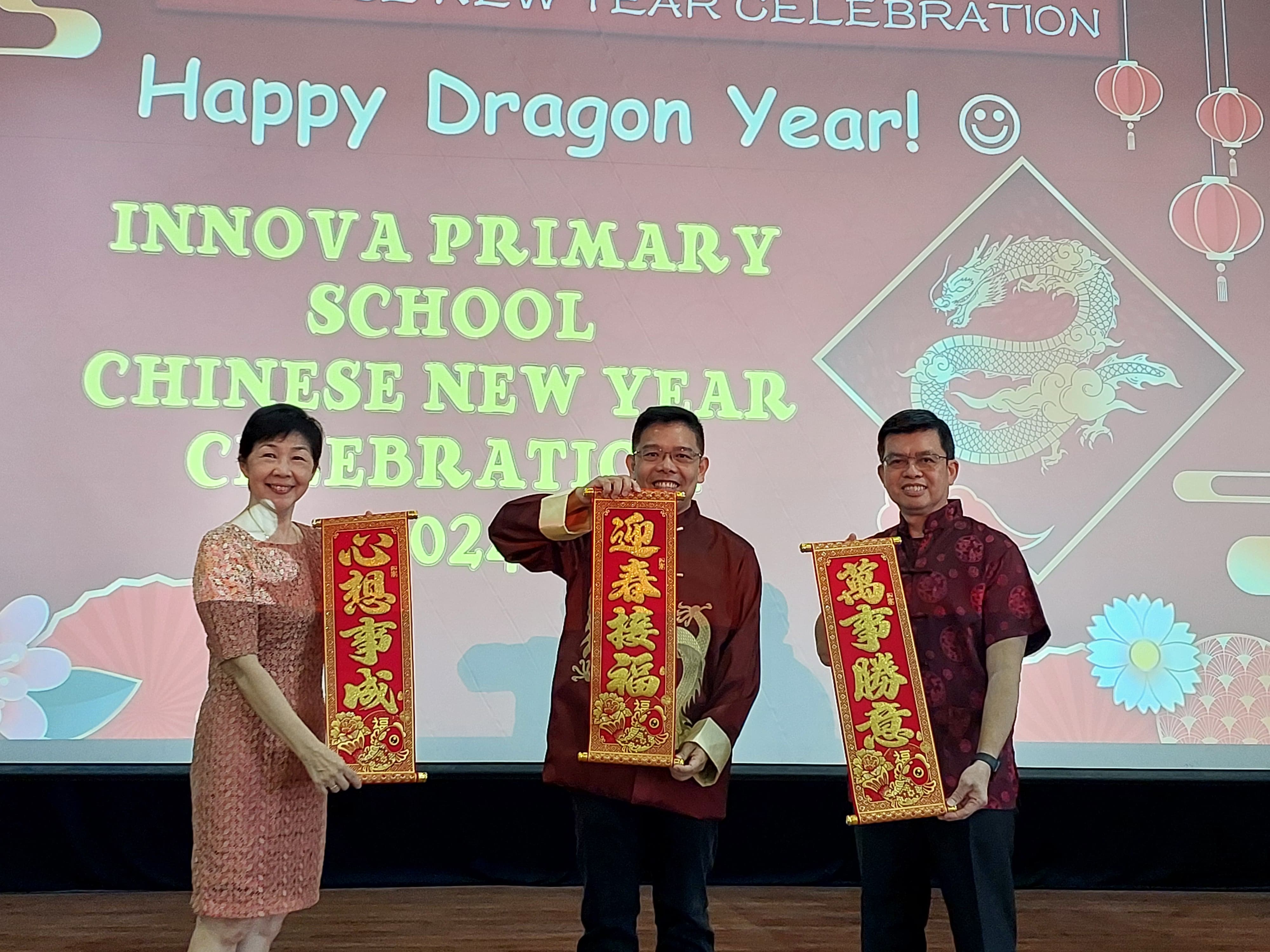Three smiling people hold red Chinese New Year scrolls at Innova Primary School's 2024 Dragon Year celebration.