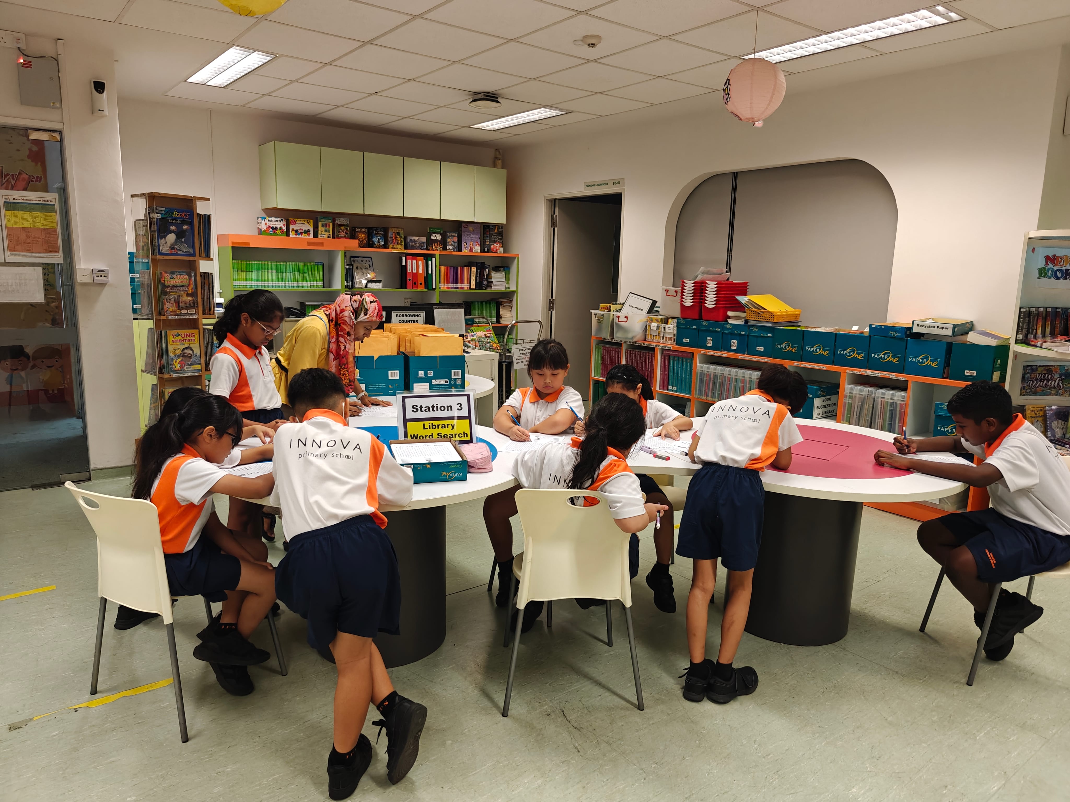 Children in school uniforms work at round tables in a library while a teacher assists.