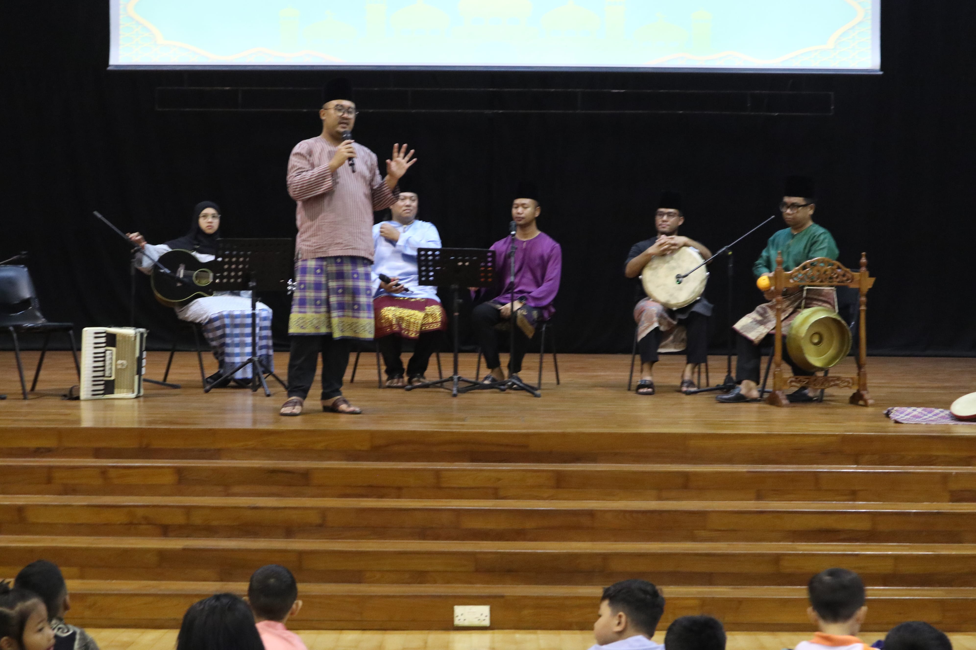 Man speaks into a microphone on stage with seated musicians holding instruments, audience children in front.