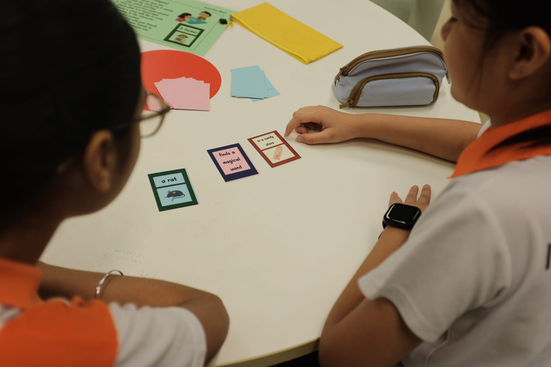Two children in school uniforms sit at a white table arranging story phrase cards reading "a rat," "finds a magical wand," and "in a candy store."