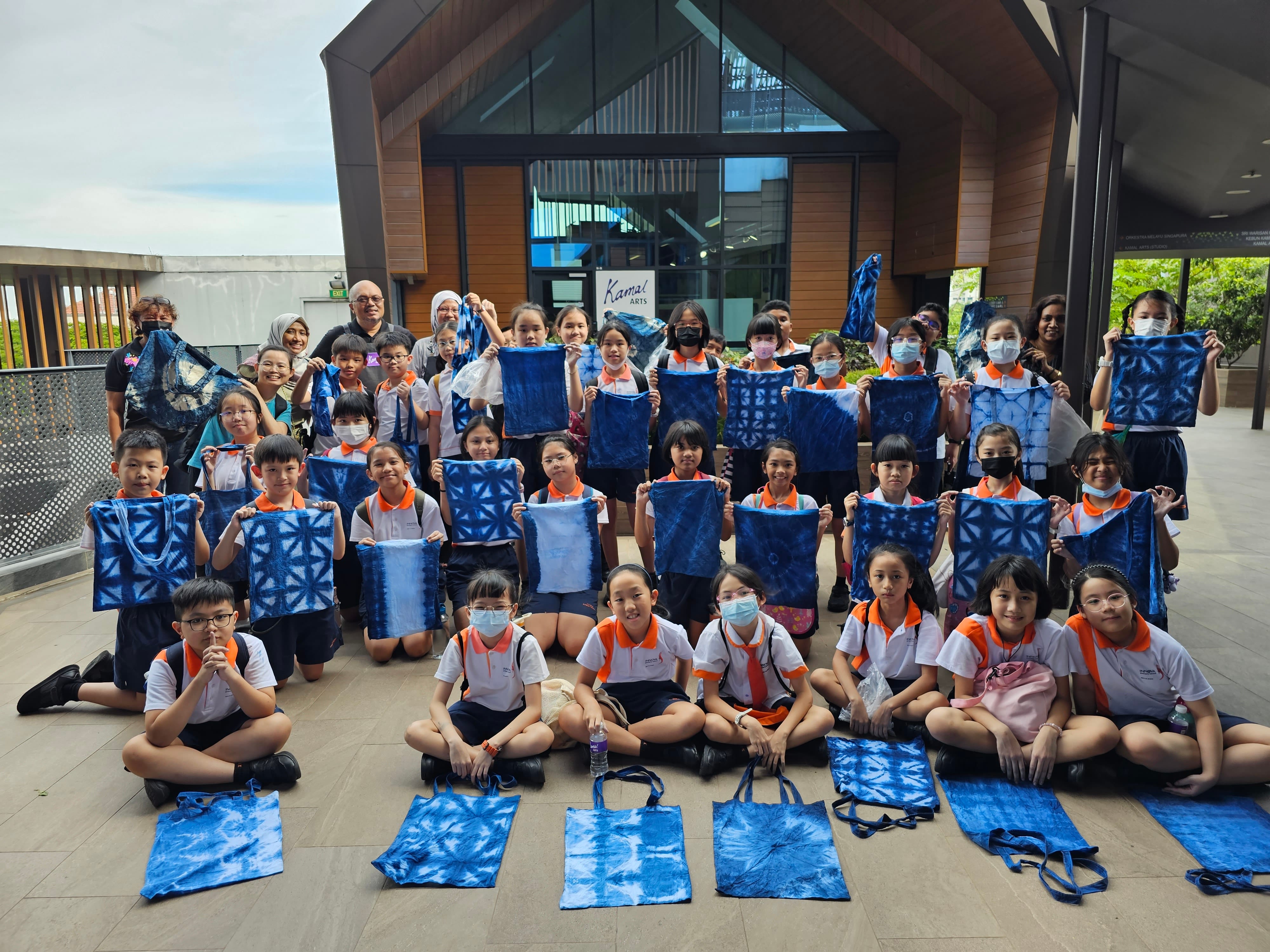 A large group of school children in uniforms hold up blue tie-dye fabric pieces and tote bags outside a building.