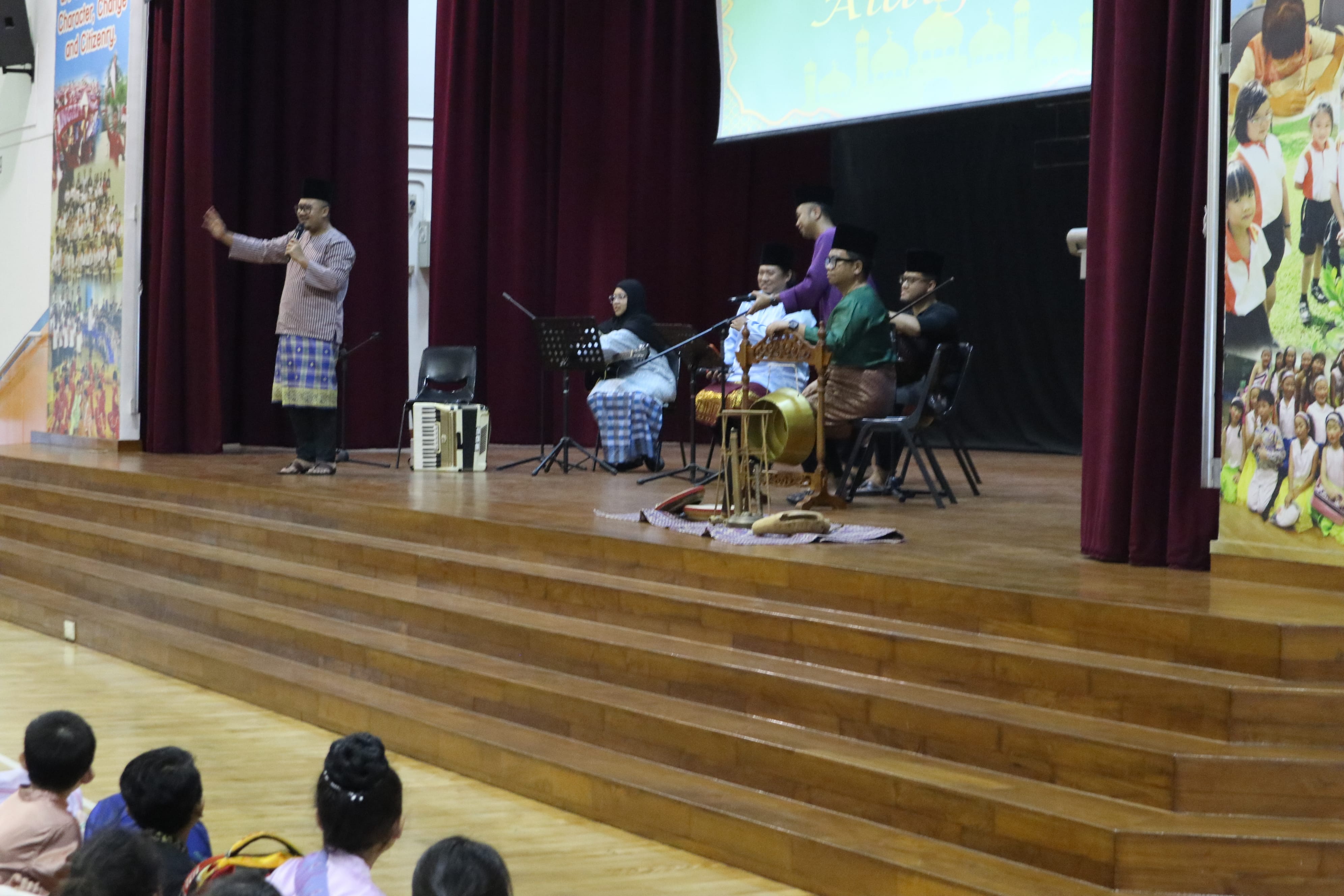 Man speaks into microphone on stage beside seated musicians and instruments, audience seated below.