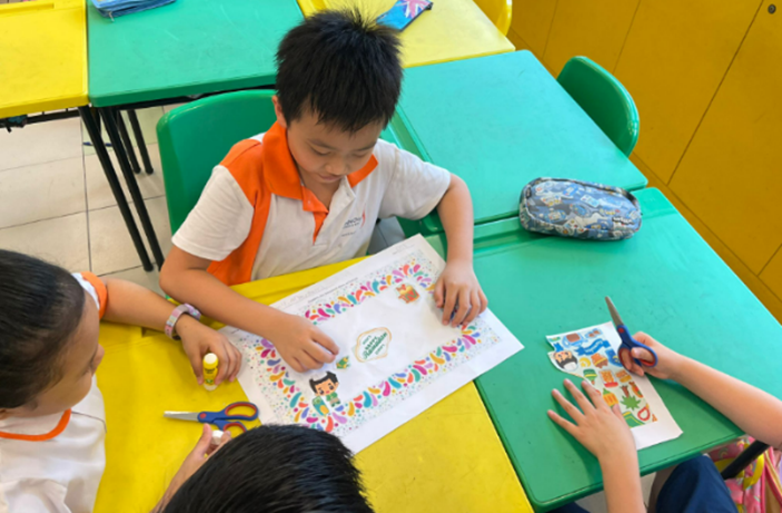 Three children sit at colorful classroom tables working on a craft activity with scissors and decorated paper.