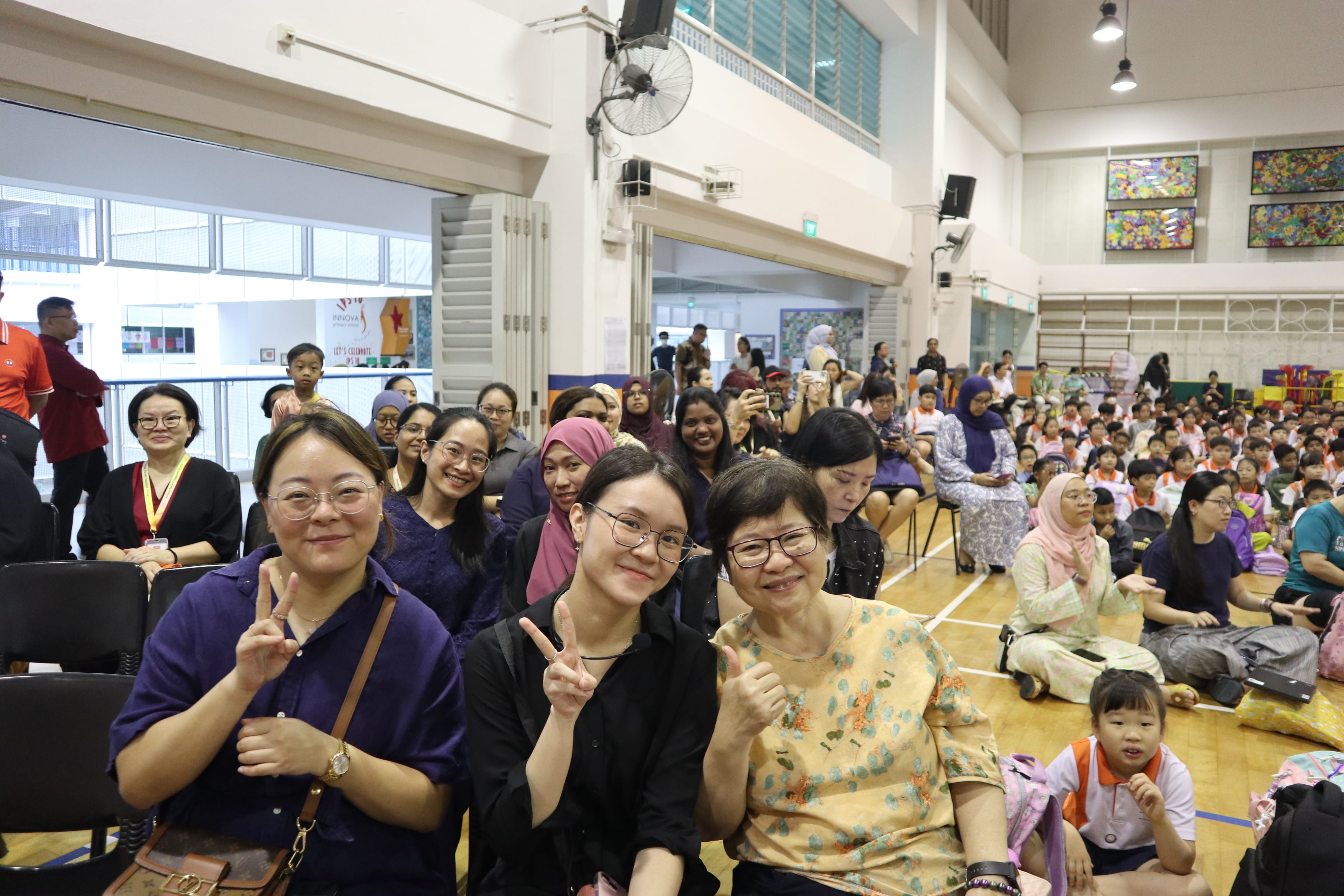 Three women smiling in front row, flashing peace signs, with a crowded school hall behind them.