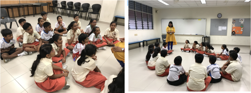 Students in uniform sit on a classroom floor in a circle while a teacher in yellow stands before a whiteboard.