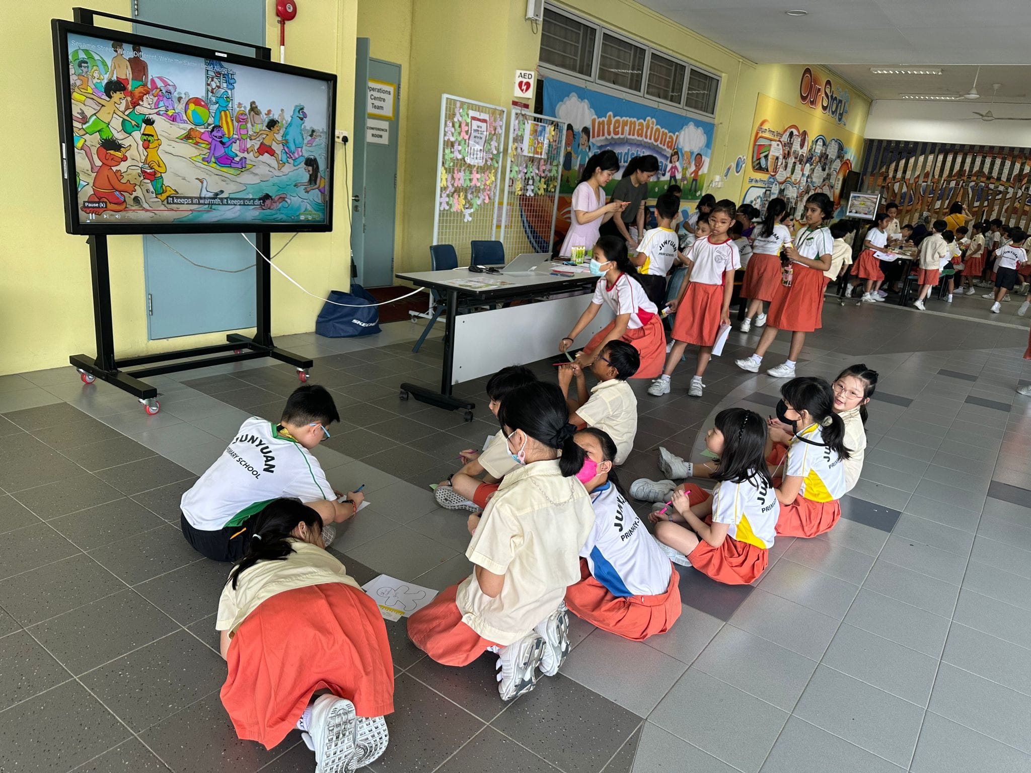 Primary school children in uniforms sit on a floor watching a Sesame Street cartoon on a large display screen indoors.