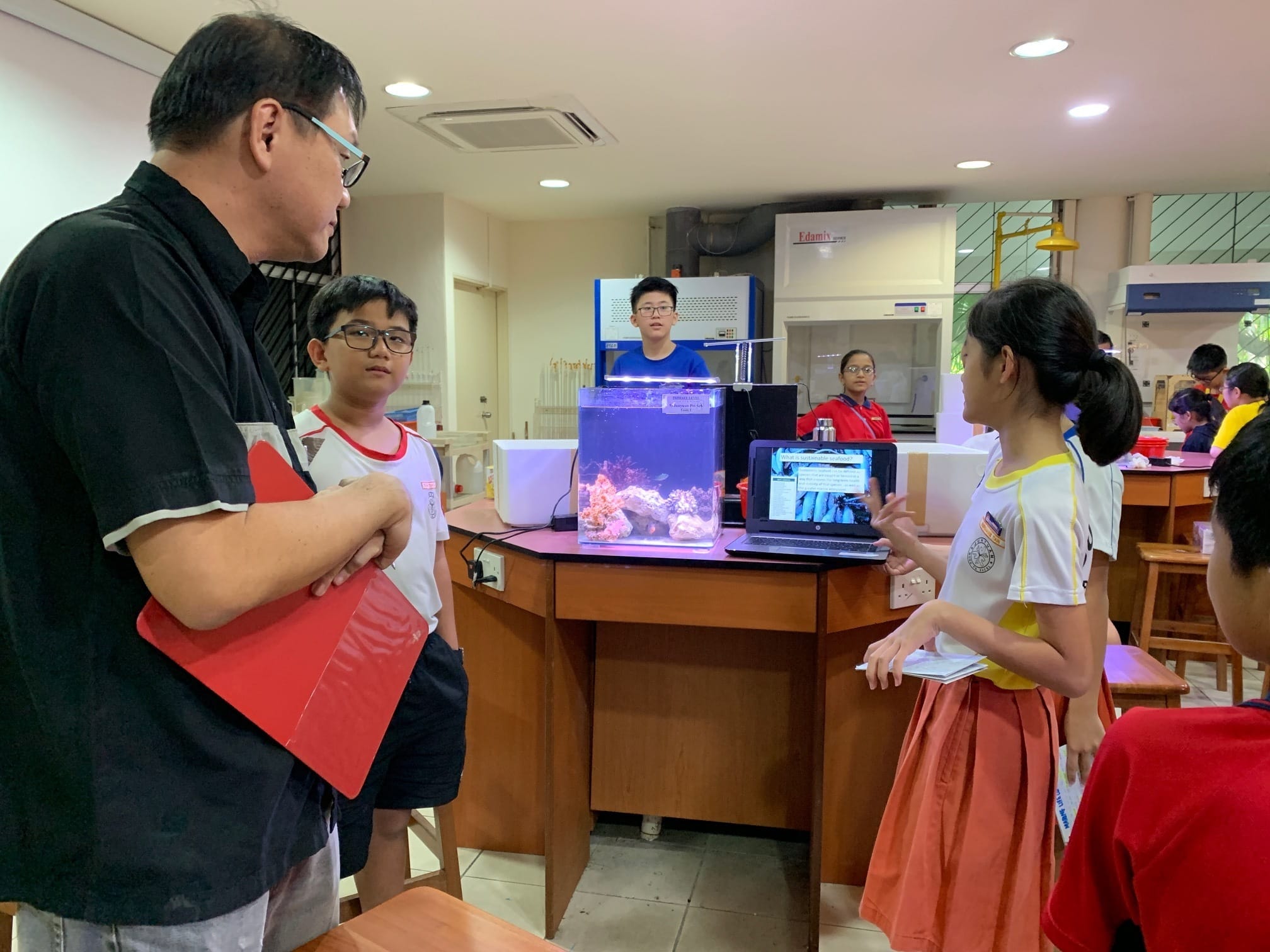 Students present a small coral reef aquarium and laptop to an adult in a school science laboratory.