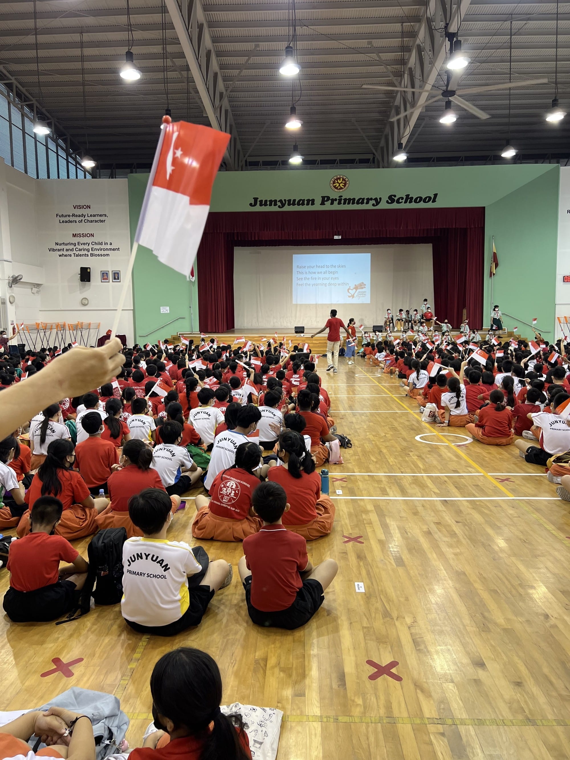 Students in red and white uniforms sit on a gym floor waving Singapore flags at a Junyuan Primary School assembly.