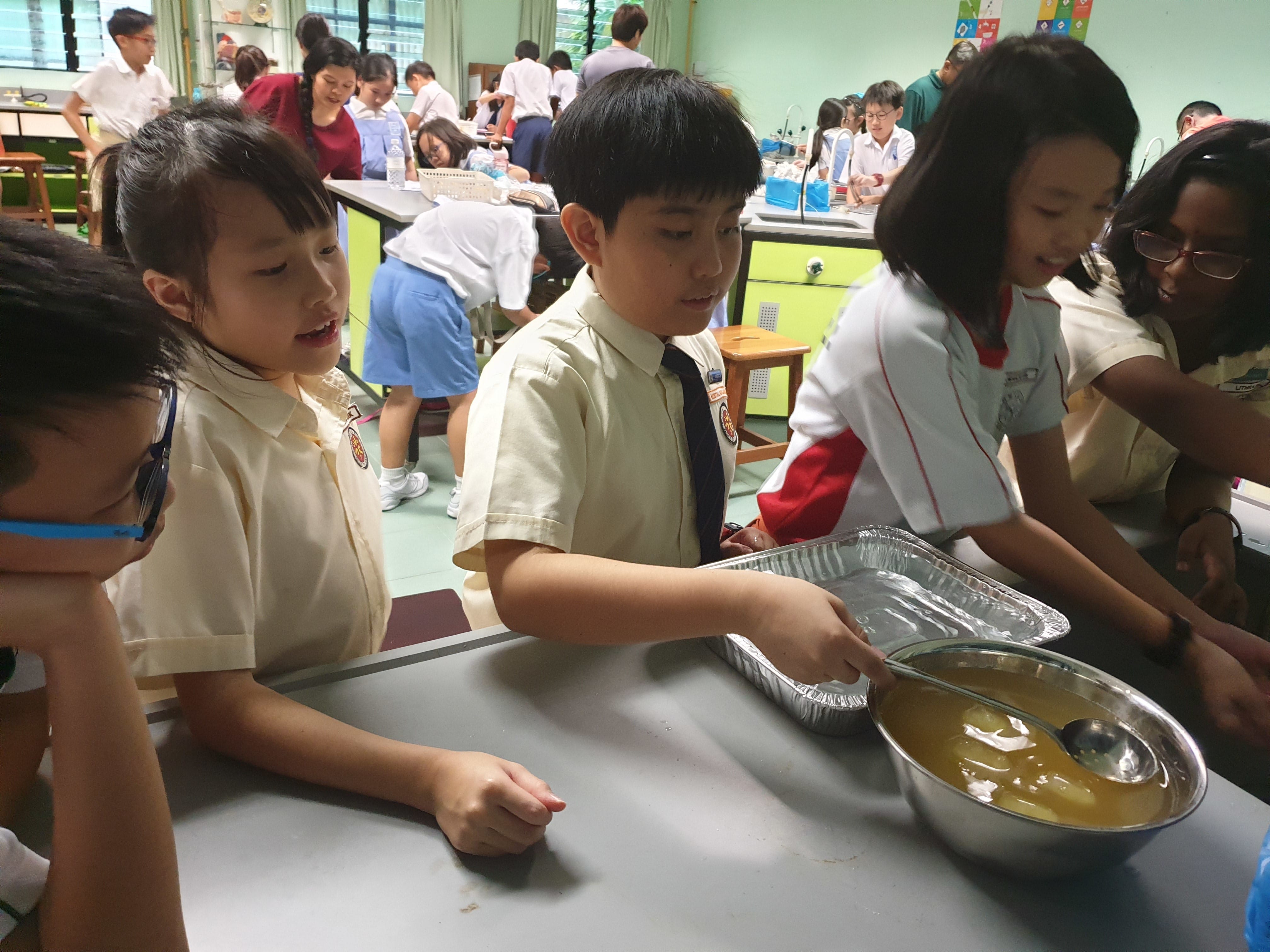 Students in school uniforms gather around a table with a bowl of brown liquid during a classroom activity.