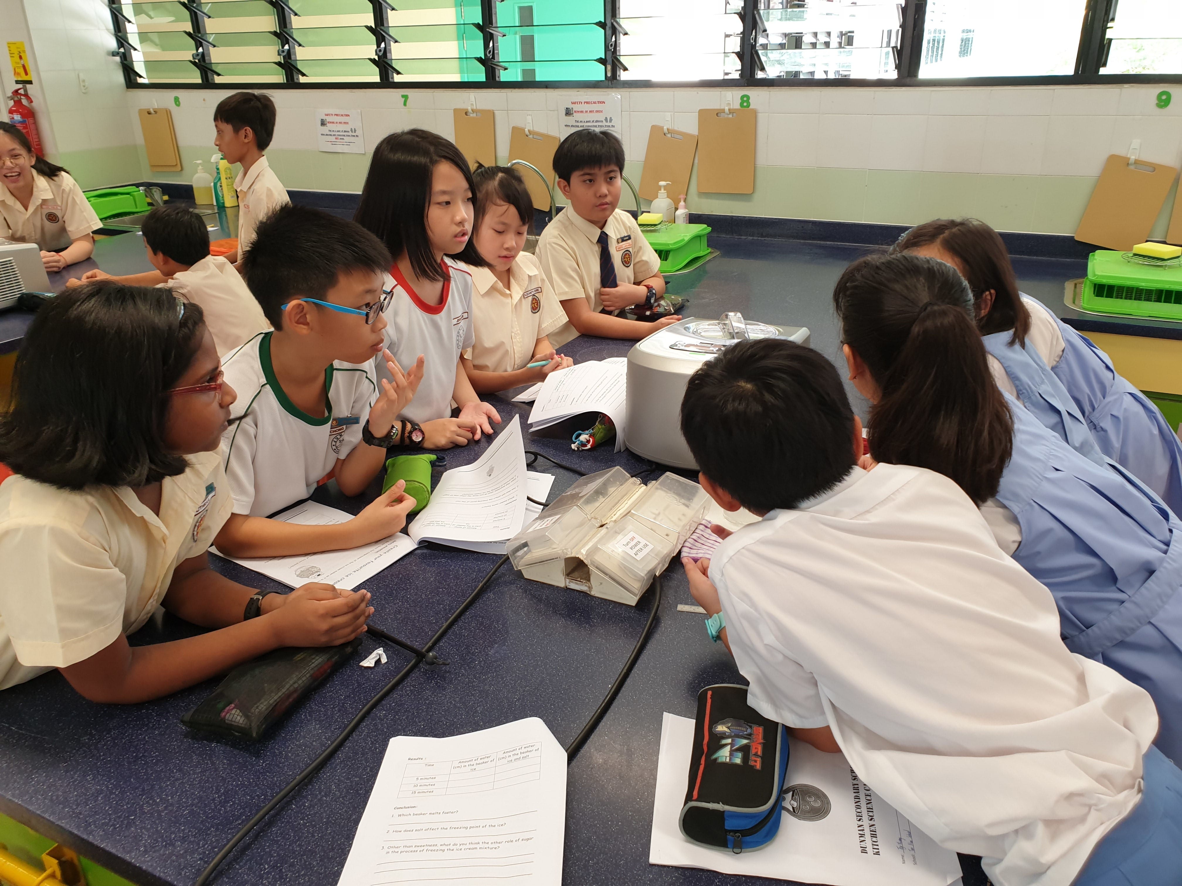 Students in uniforms gather around a blue lab table with worksheets and equipment during a science class.
