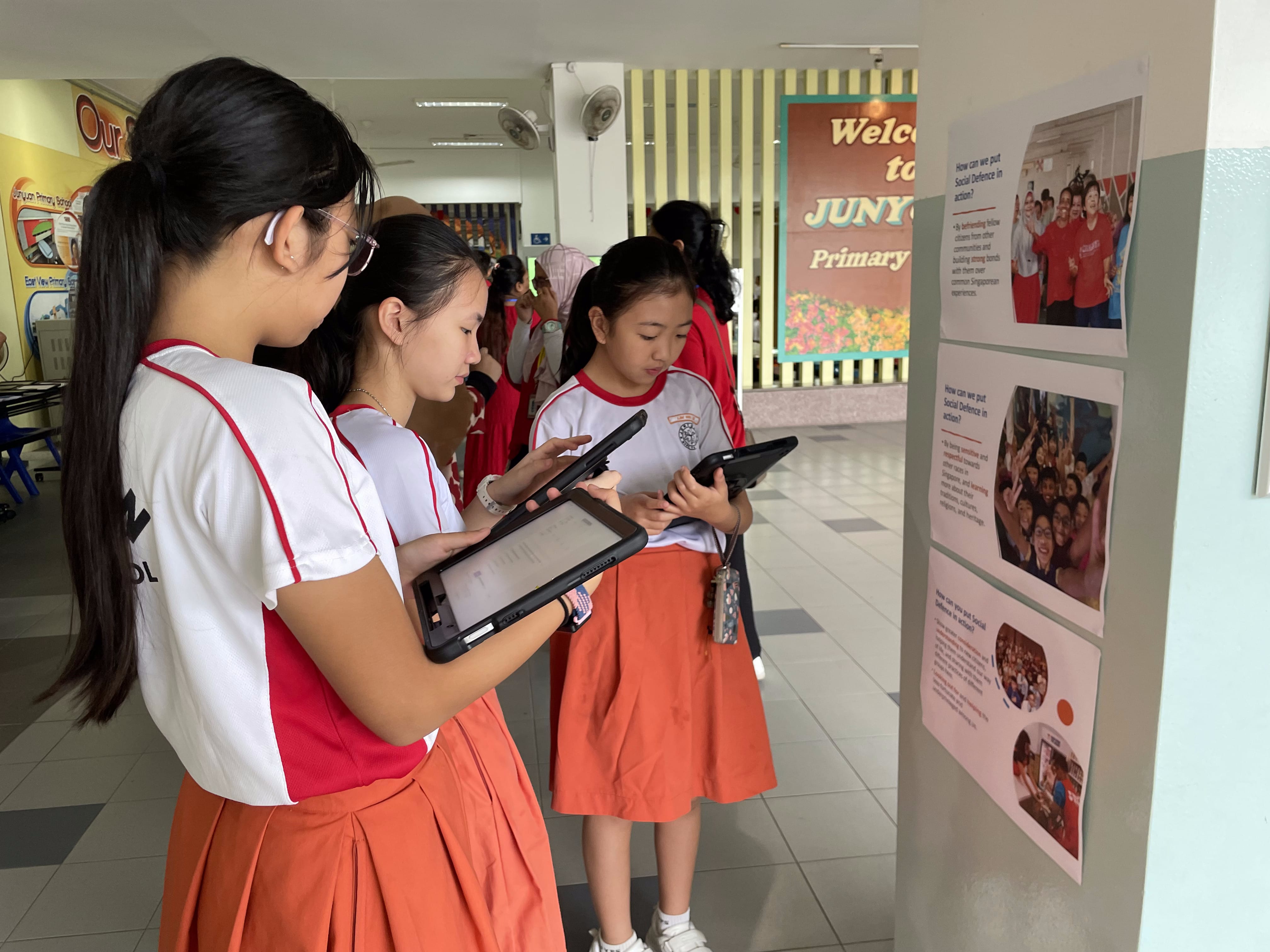 Three schoolgirls in red and white uniforms hold tablets while viewing a display board in a school corridor.