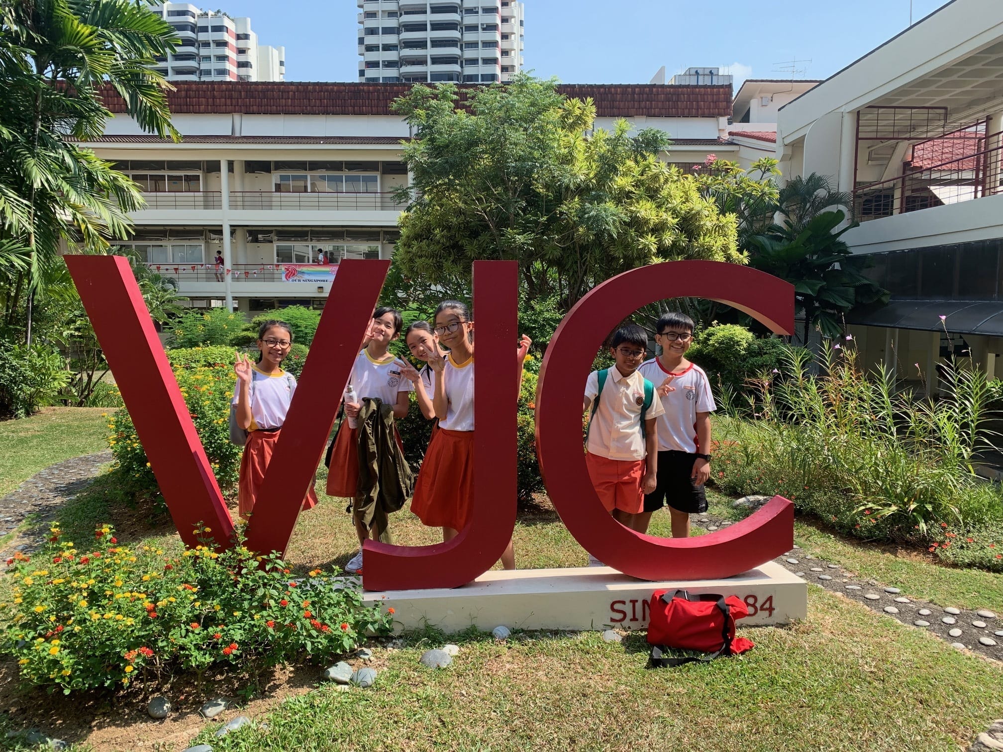 Five uniformed students pose with peace signs beside a large red VJC sign on a sunny school campus.