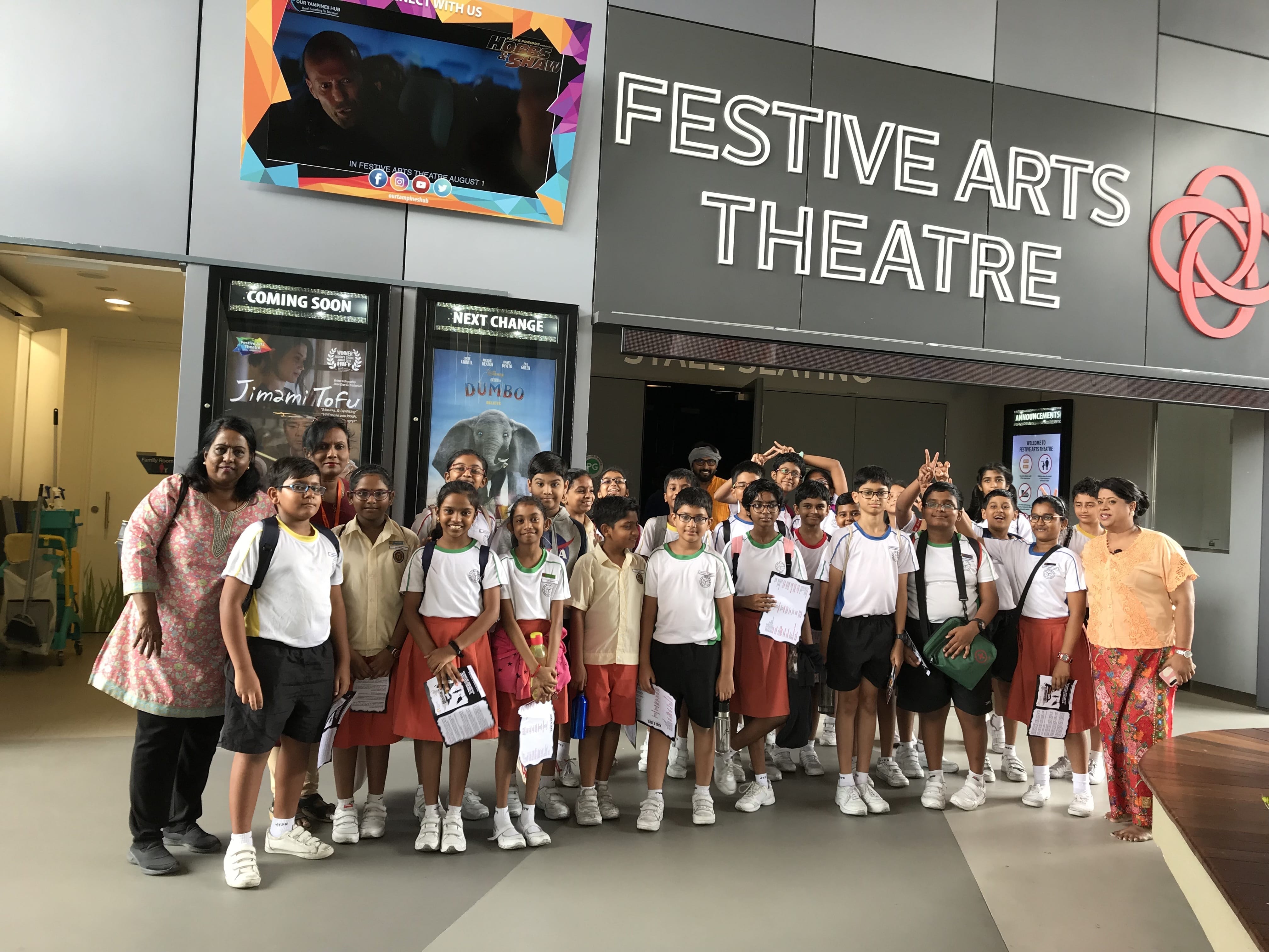 A group of school children and two adults pose in front of the Festive Arts Theatre entrance.