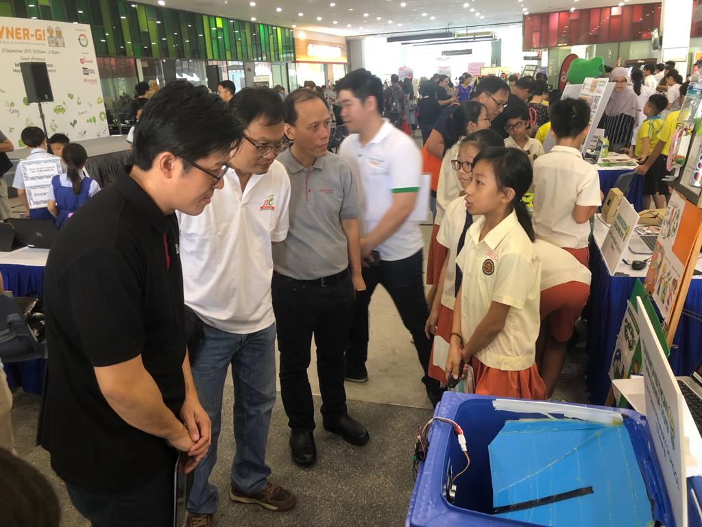 Students and adults gather at an indoor exhibition fair with display booths and project boards.