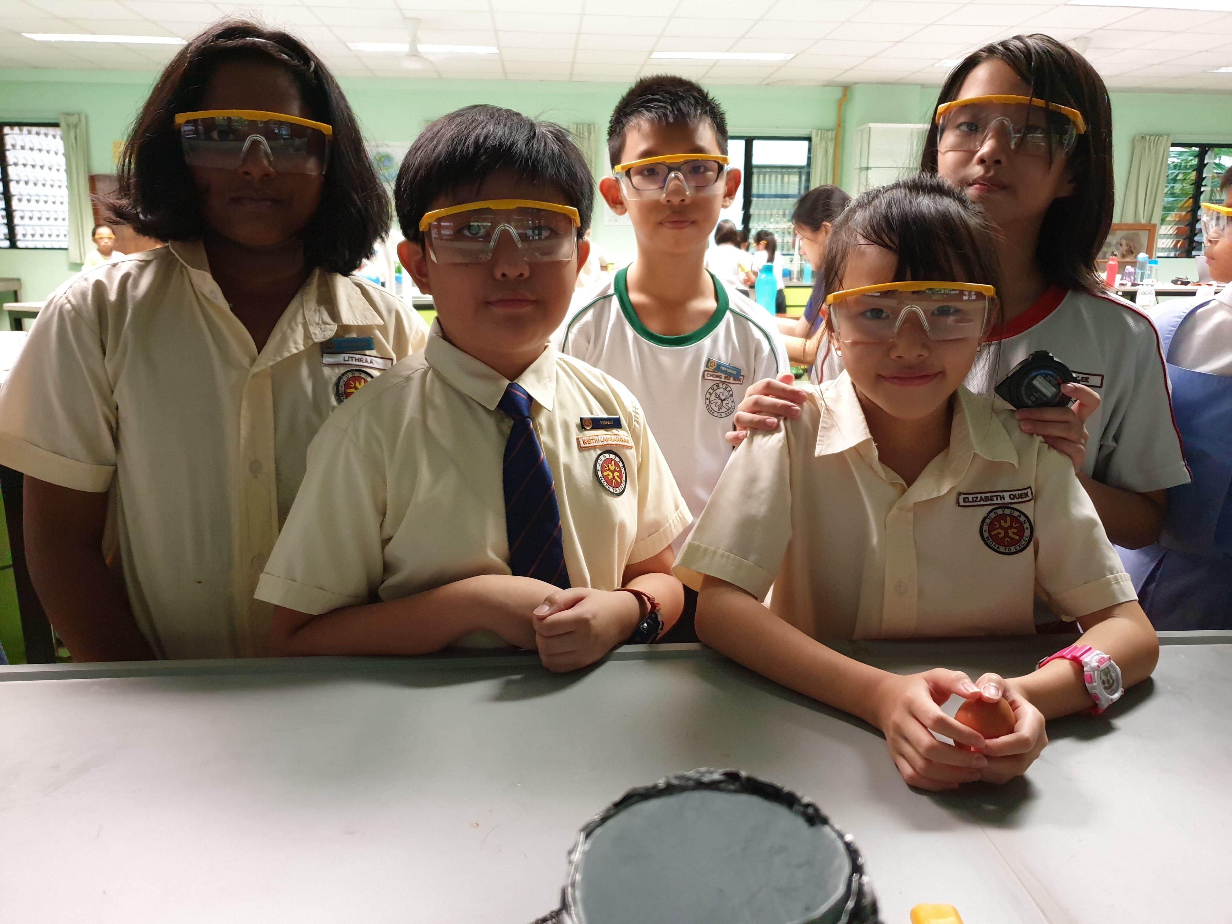 Five school children in uniforms wearing yellow-framed safety goggles stand at a lab table in a science classroom.