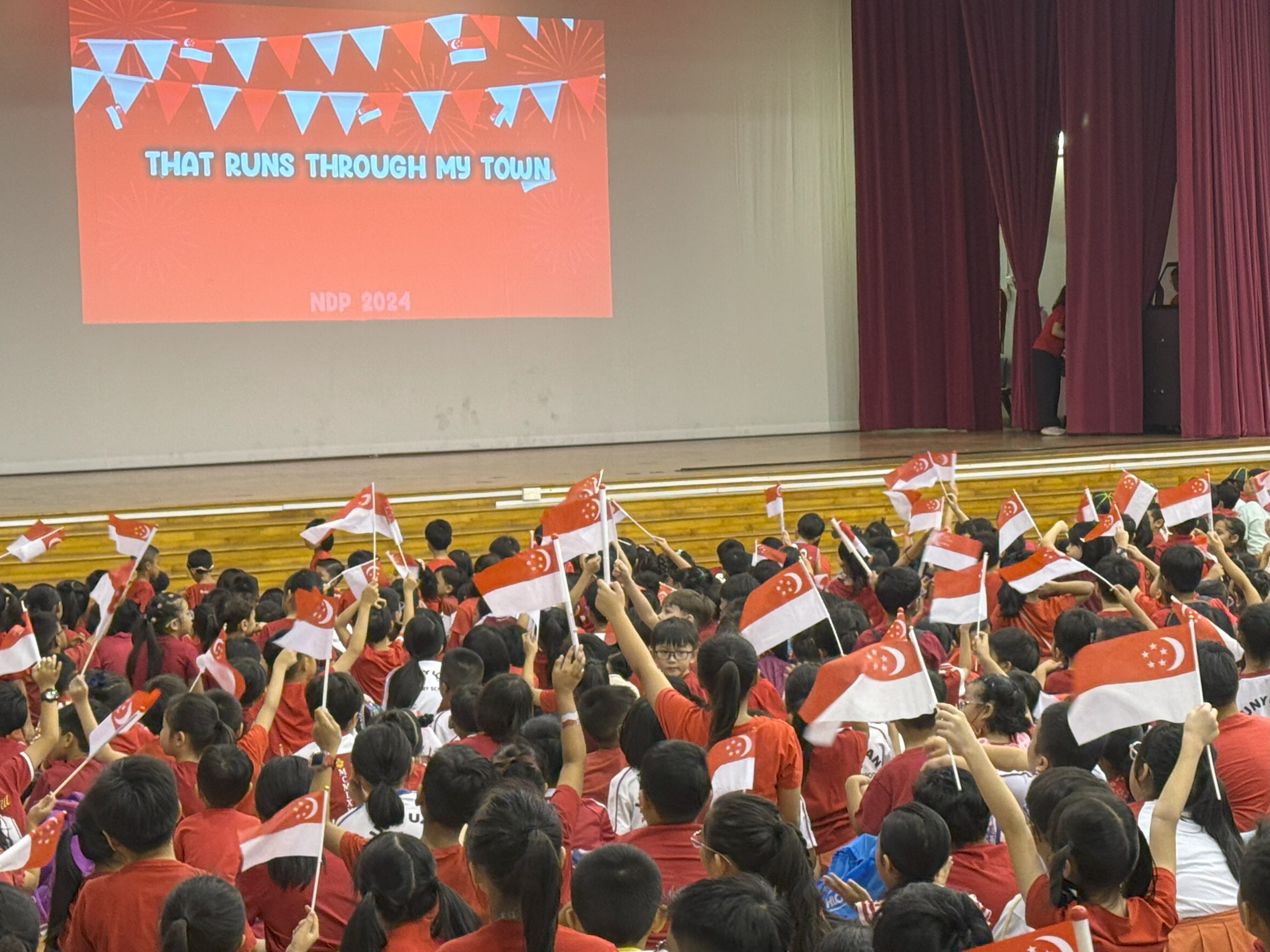 Children in red shirts wave Singapore flags at an NDP 2024 school event with a festive screen displayed.