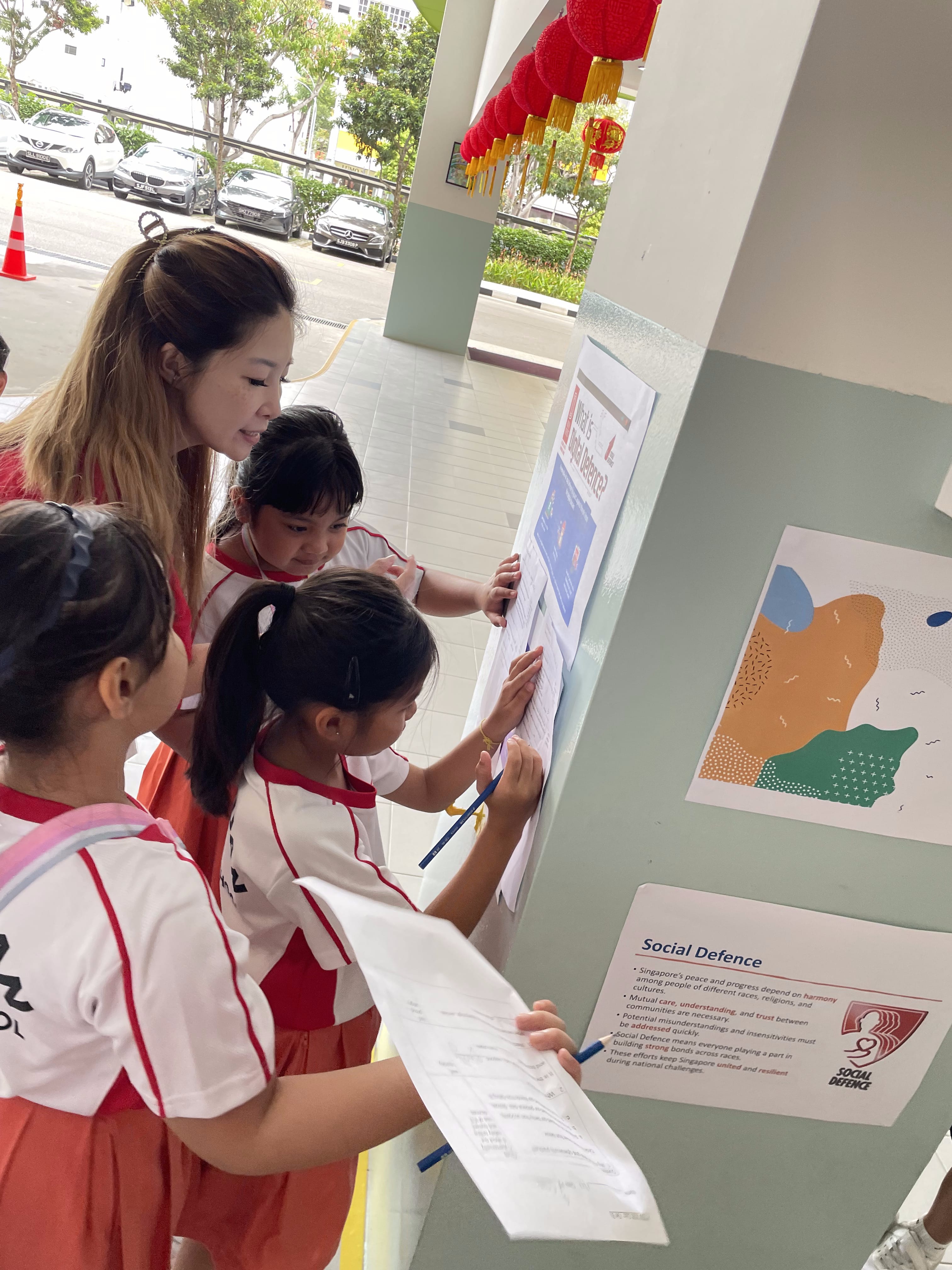 A teacher and three young students in school uniforms read a Social Defence display board outdoors.