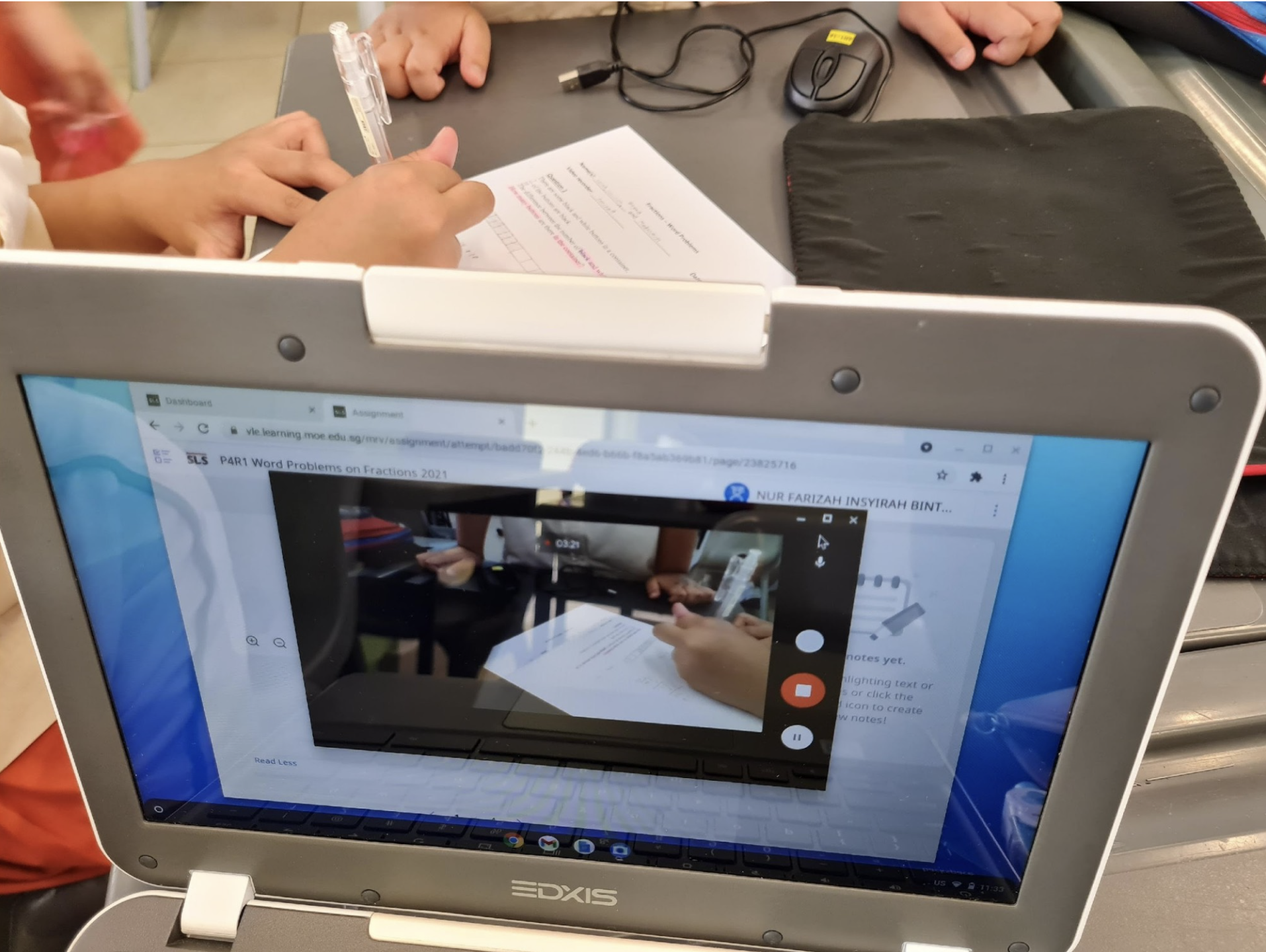 Student holds pen over worksheet beside an EDXIS laptop showing a math fractions assignment with a video playing.