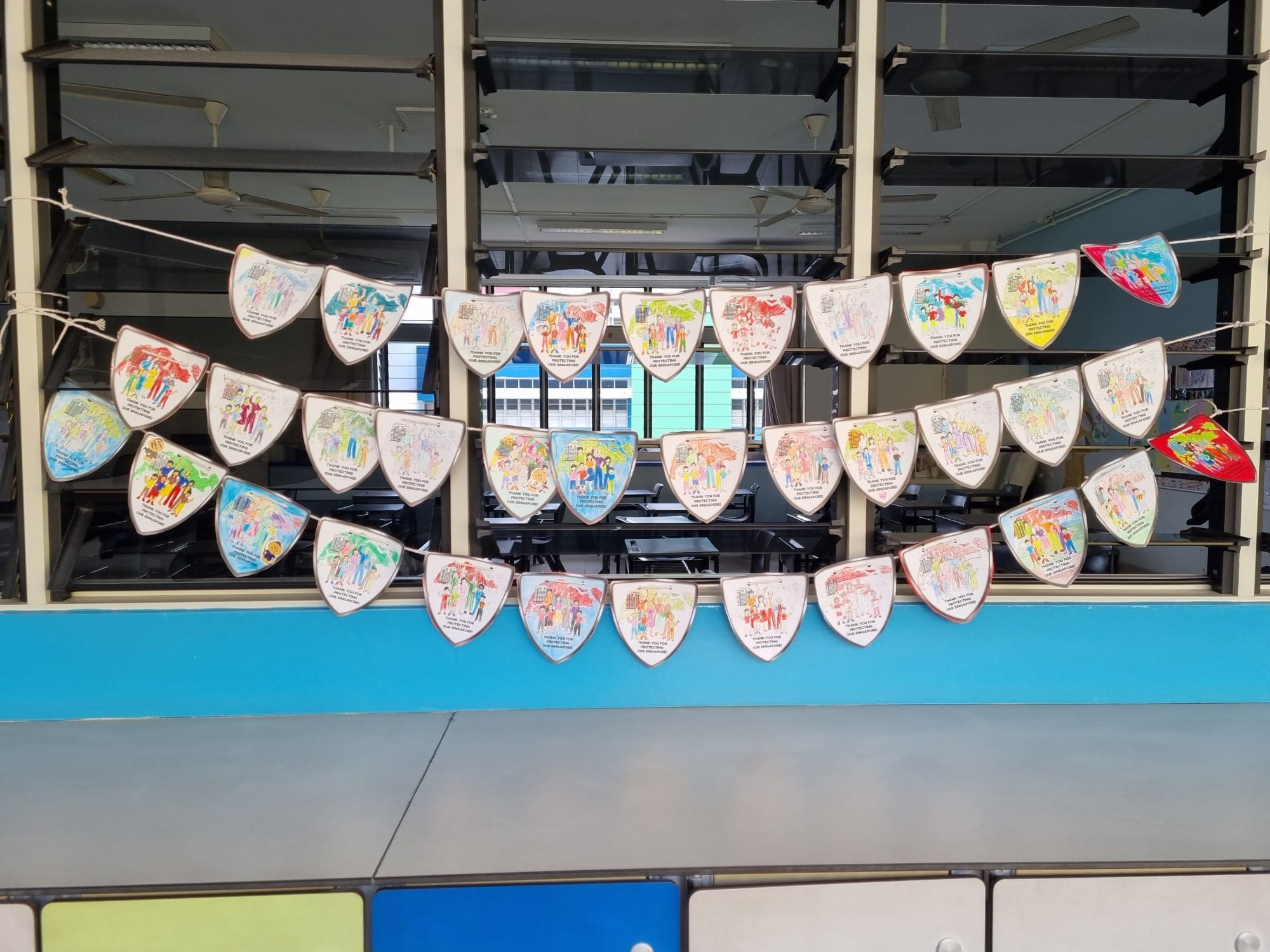 Colorful hand-drawn shield-shaped paper bunting with people illustrations strung in three rows indoors.