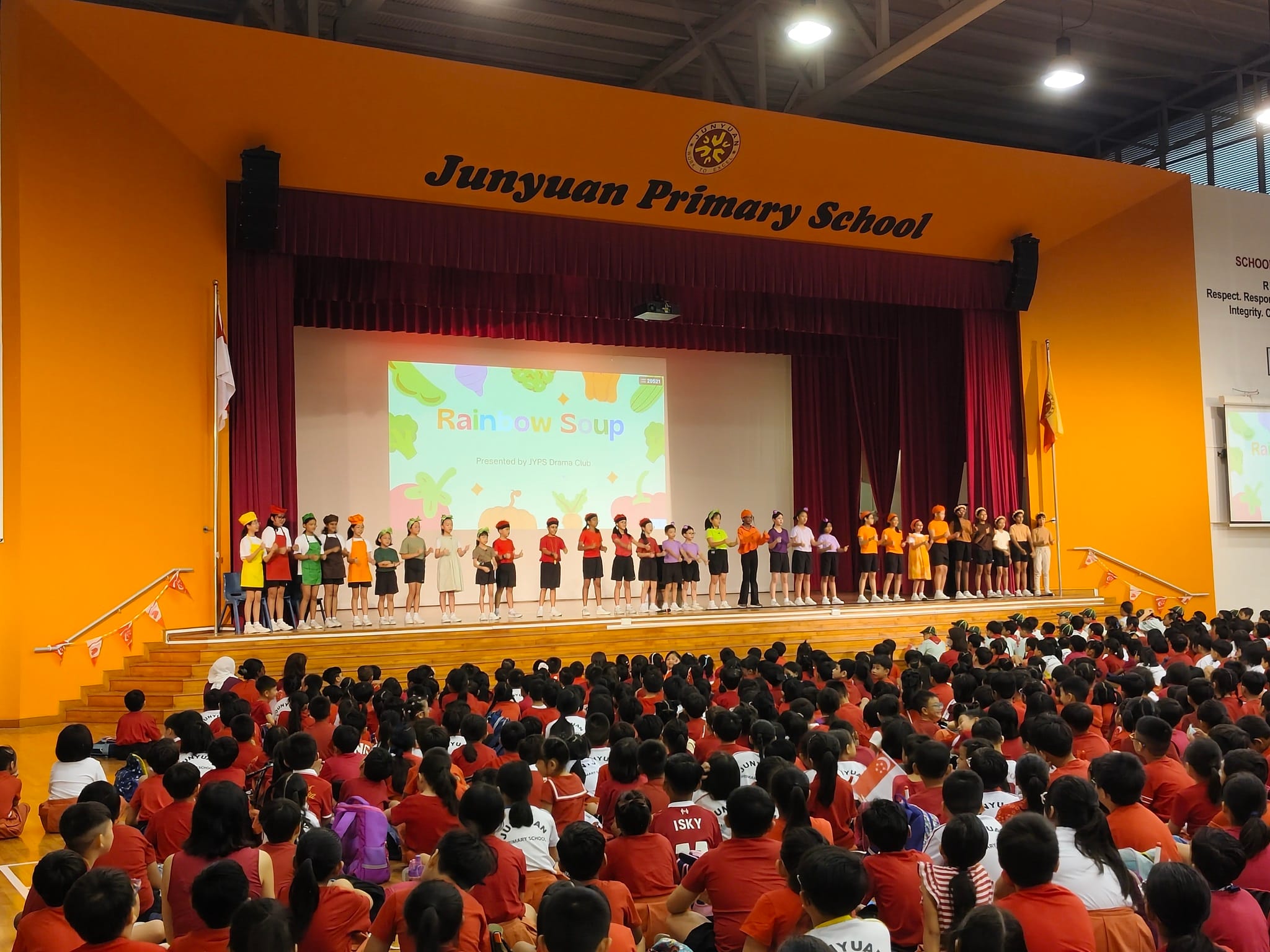 Students in colorful costumes perform Rainbow Soup on stage at Junyuan Primary School before a large seated audience.
