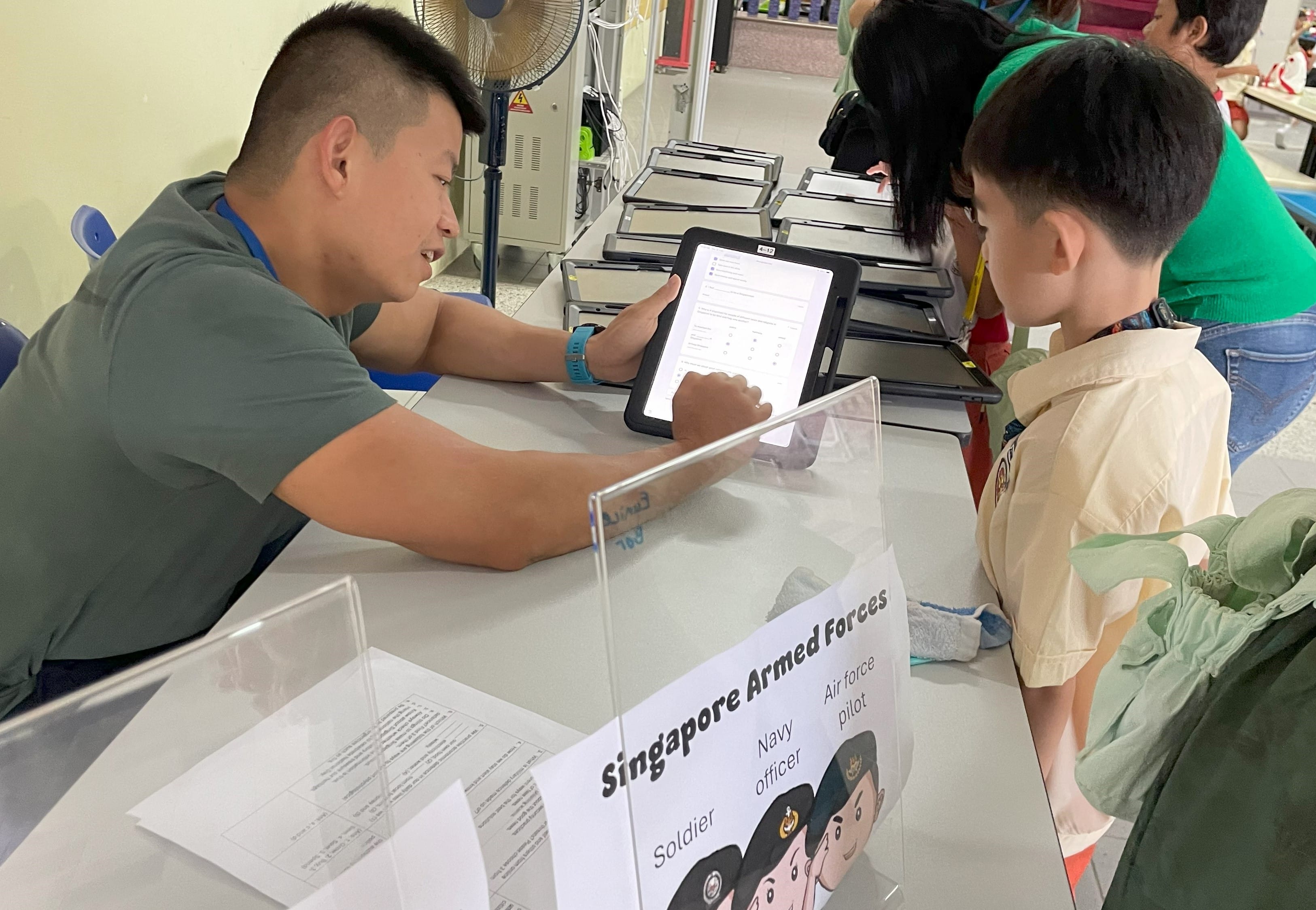 A young man shows a tablet to a child at a table with a Singapore Armed Forces sign displaying soldier roles.