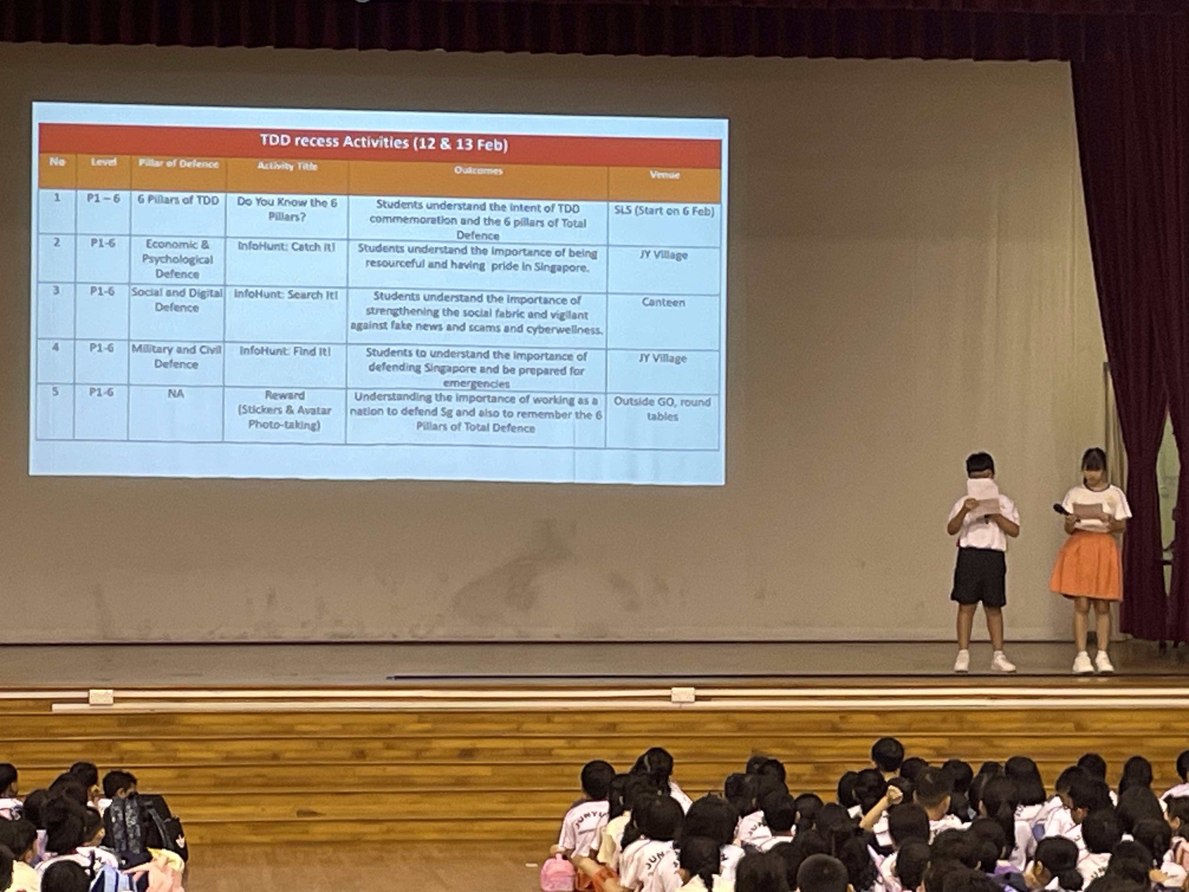Two students present on stage before a seated audience, with a projected slide showing TDD recess activities for Feb 12 and 13.
