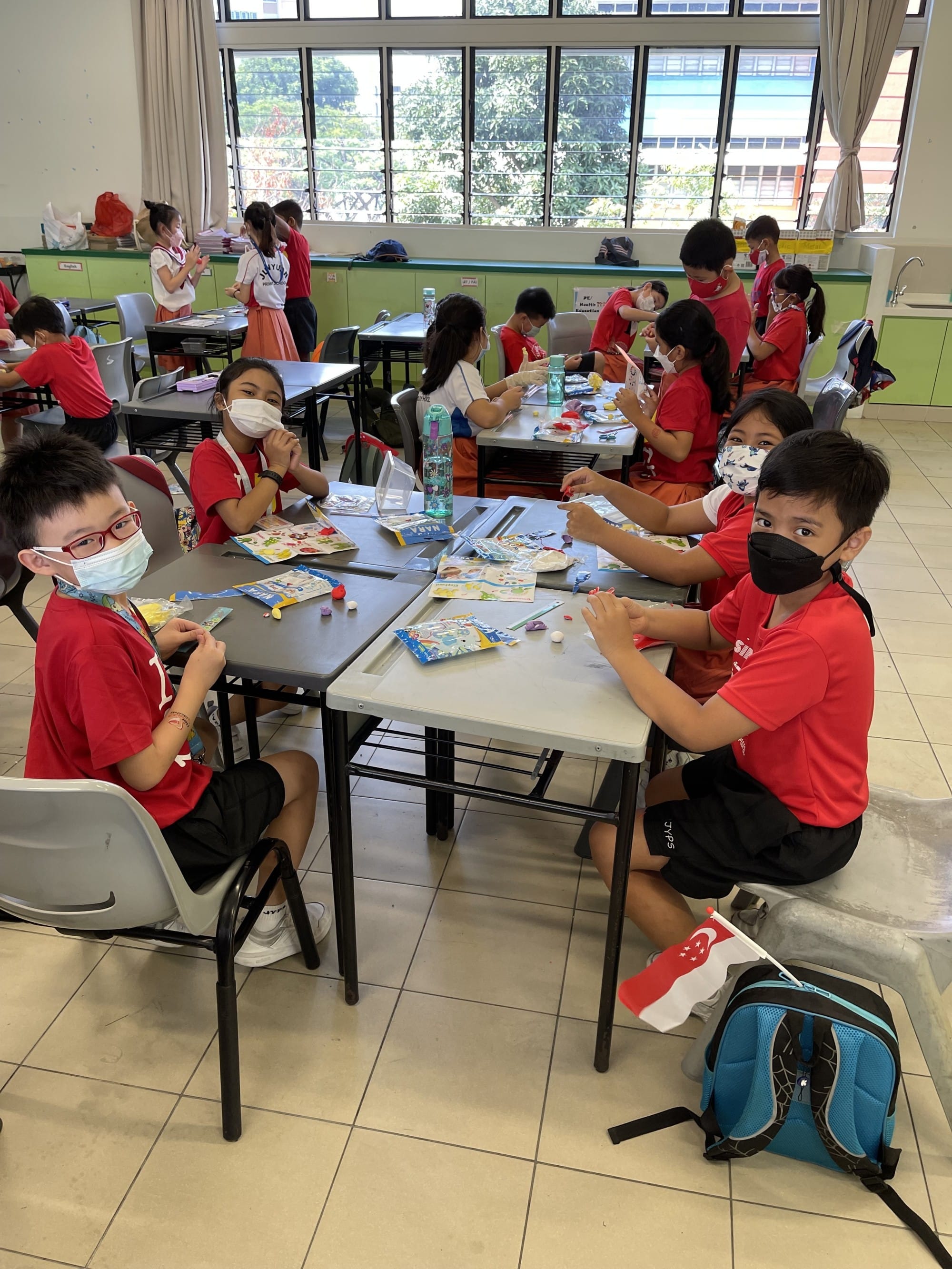 Children in red shirts and face masks sit at classroom tables doing a craft activity with colorful materials.