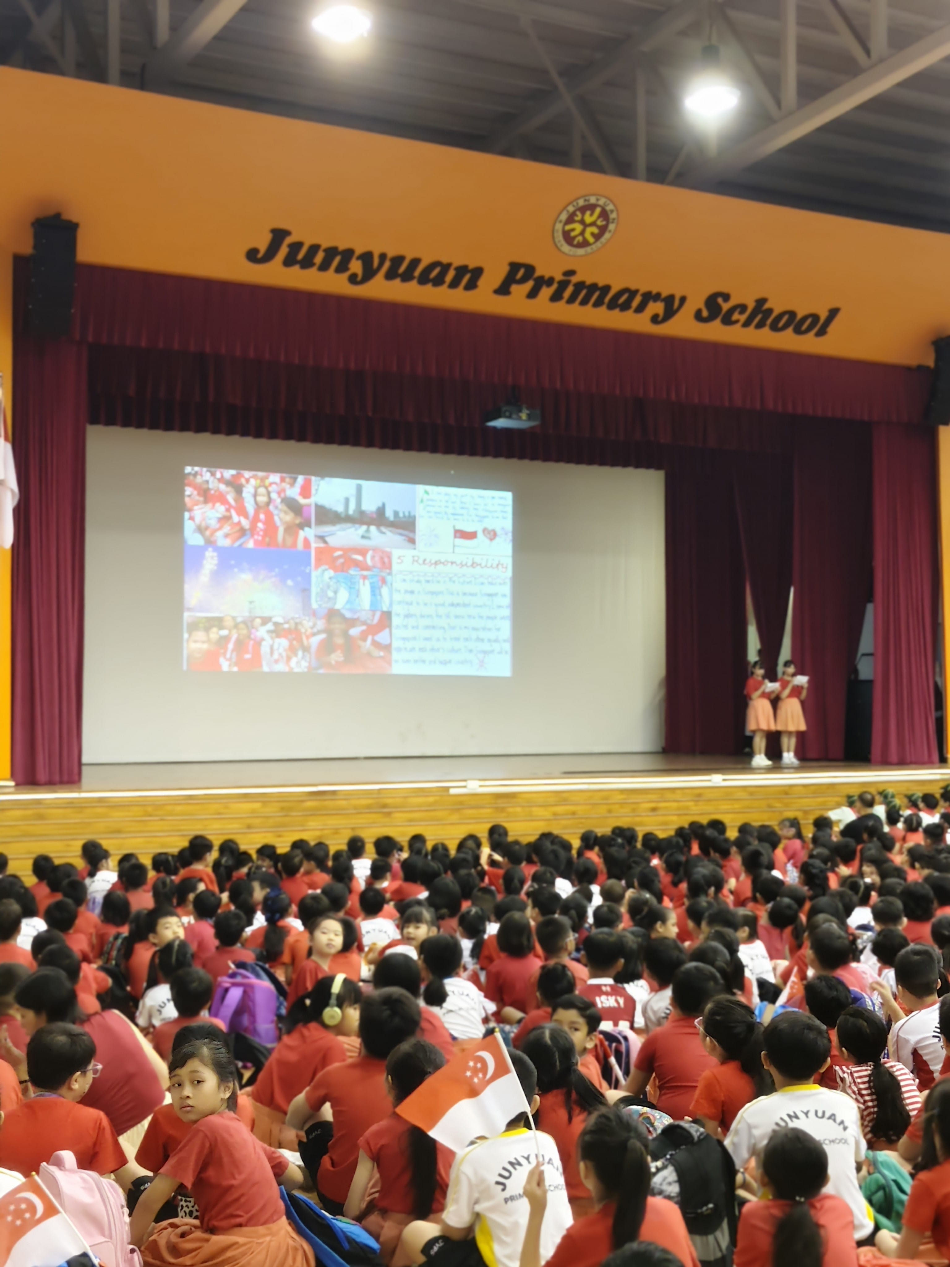 Students in red sit on a hall floor at Junyuan Primary School, facing a stage with a projected presentation.
