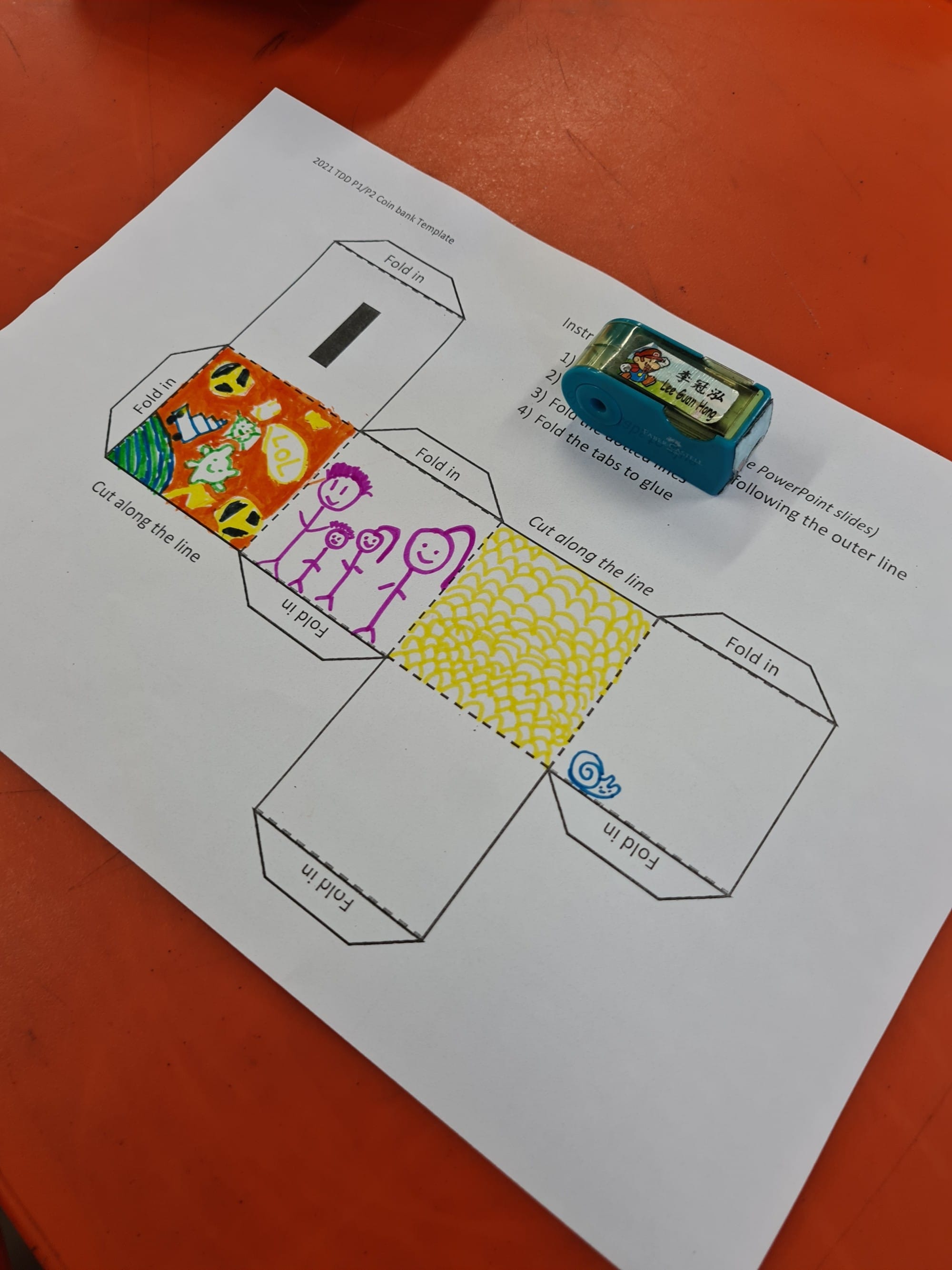 A coin bank paper craft template with colorful hand-drawn designs and a blue pencil sharpener on an orange desk.